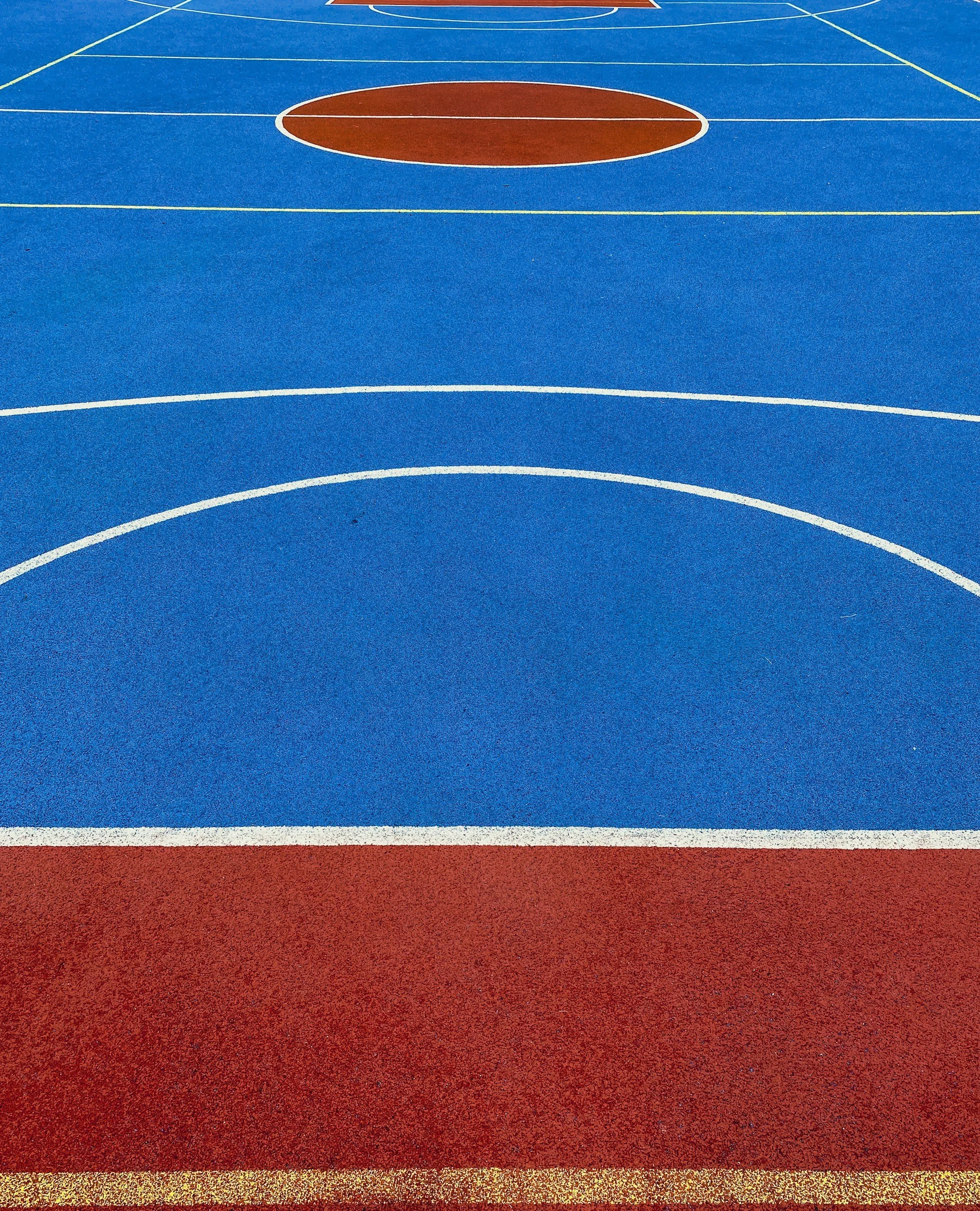 Colorful outdoor basketball court with blue and red surface, white boundary and court lines, and a large brown circle at the center.