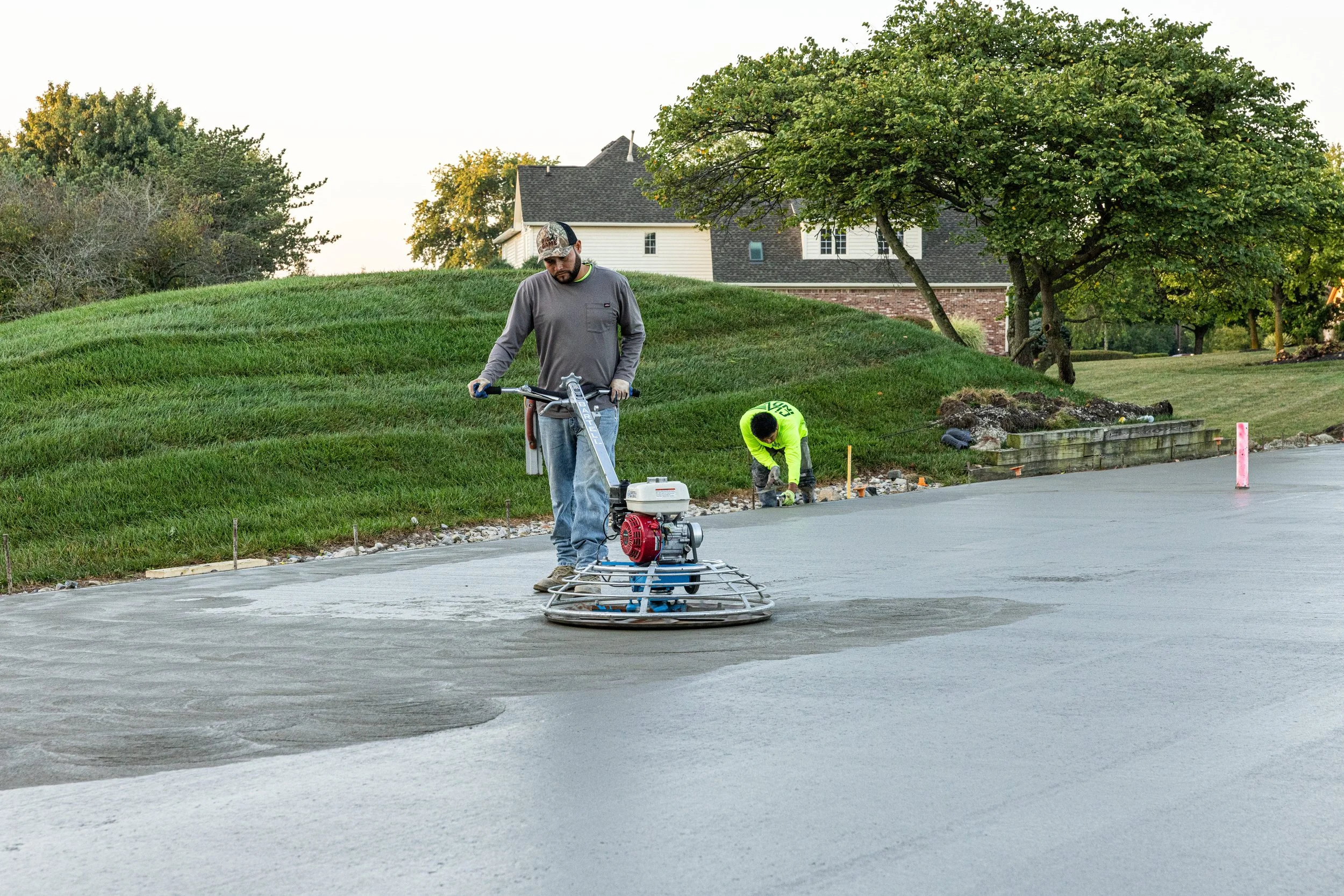 Workers paving a concrete road with one person operating a power trowel and another working nearby with tools, in a residential area with grassy hills, trees, and houses in the background.