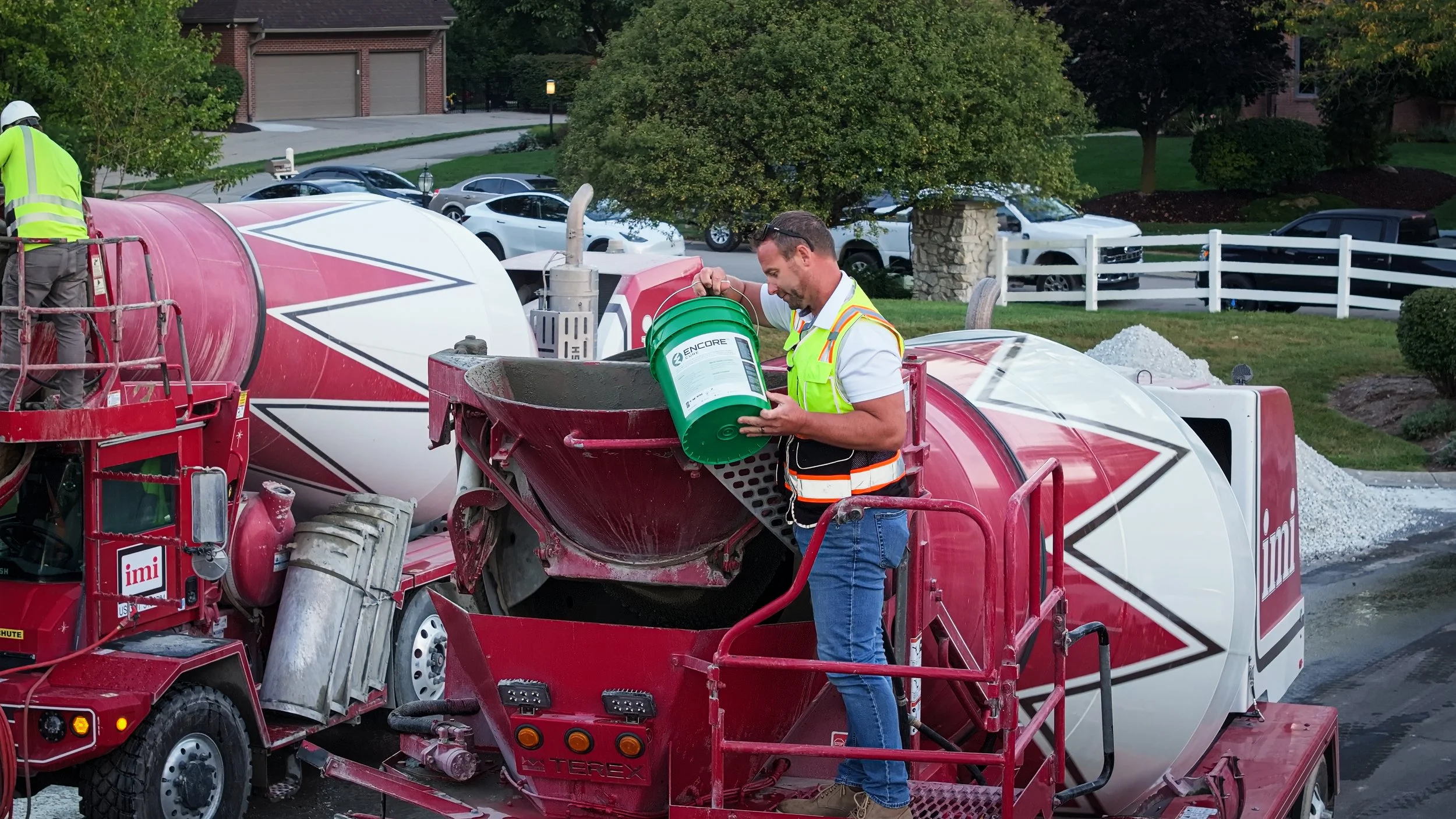 Construction worker in a neon safety vest pouring concrete from a green bucket into a cement mixer on a residential street with parked cars, trees, and houses in the background.