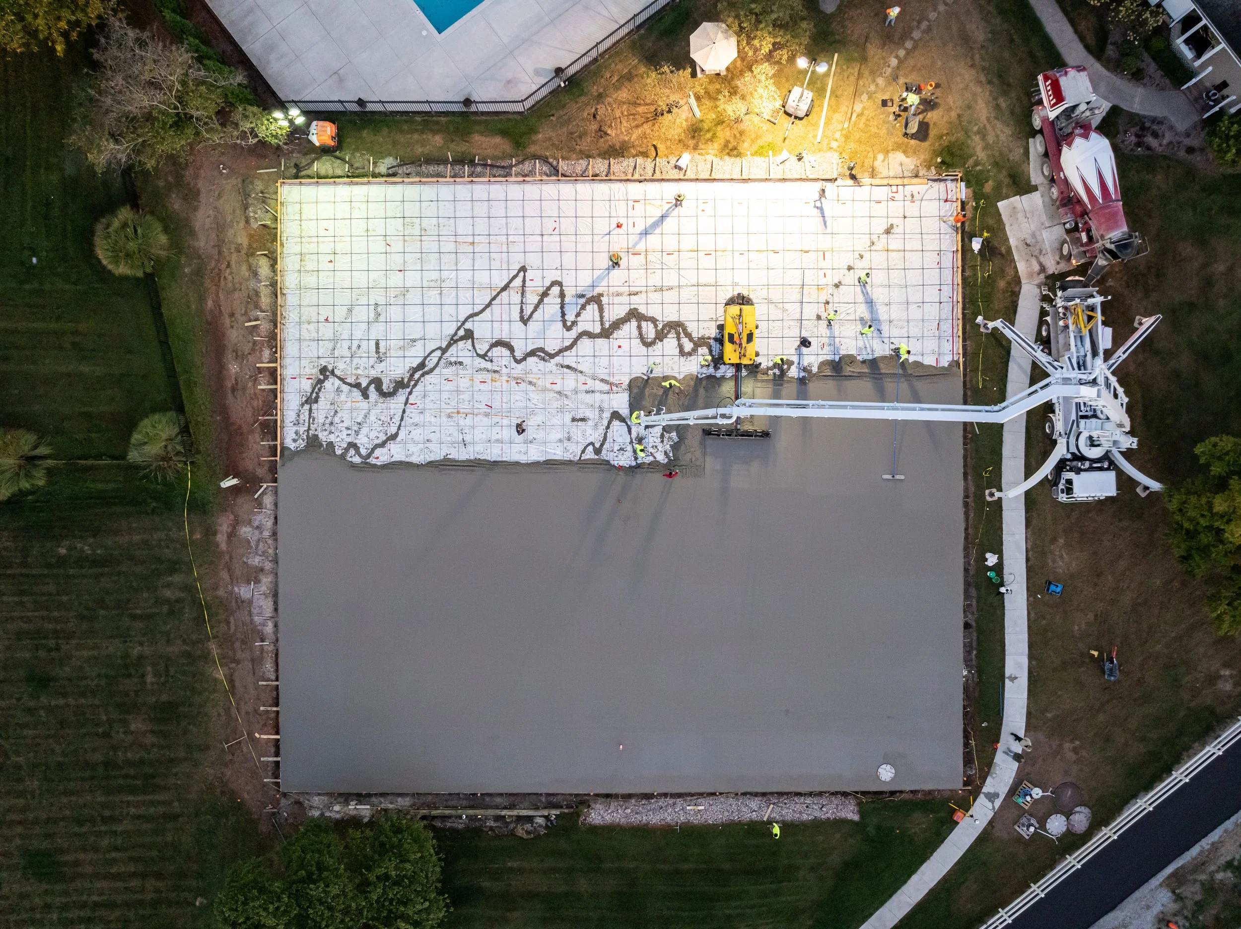 Aerial view of a construction site where workers are pouring and leveling concrete on a large, rectangular foundation. The foundation has a black line drawing of a fluctuating line graph on it. Construction equipment, including a crane and concrete t