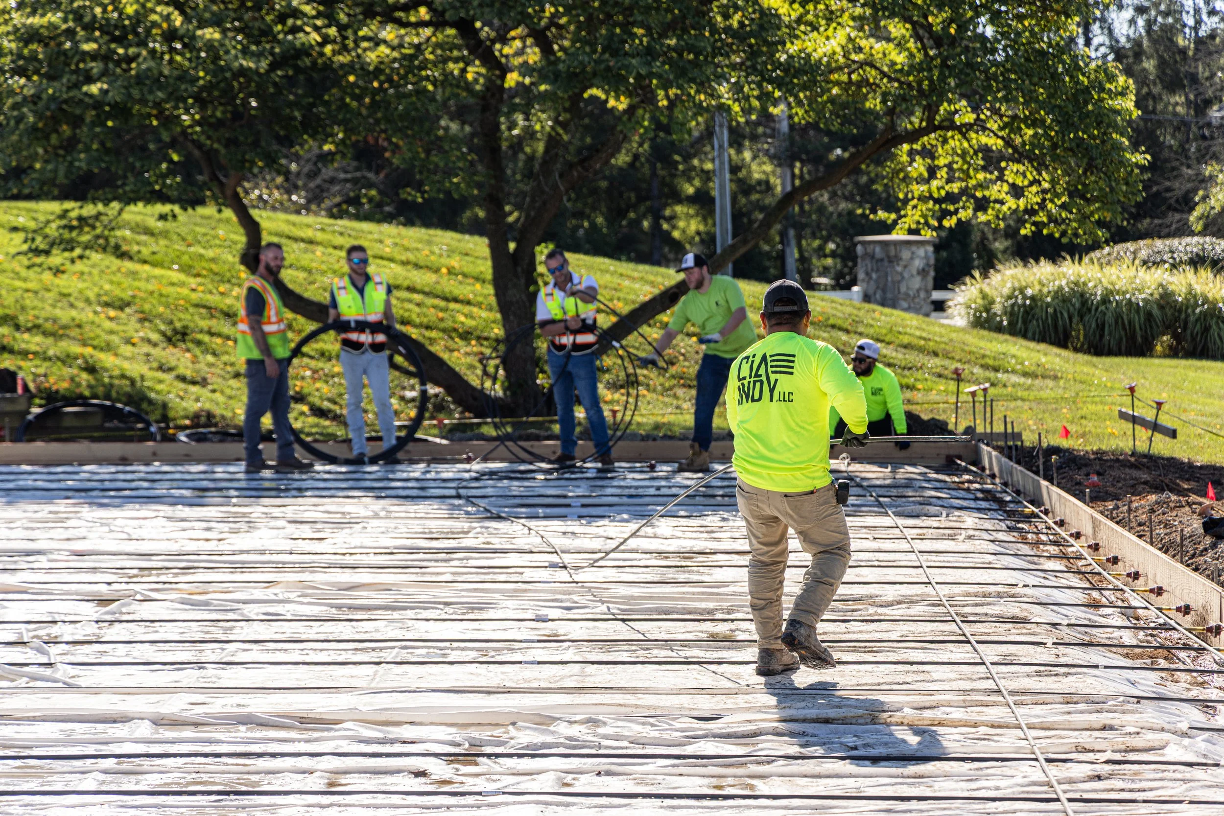 Construction workers in bright yellow and orange safety vests working on a concrete slab foundation outdoors, with a grassy hill and trees in the background.