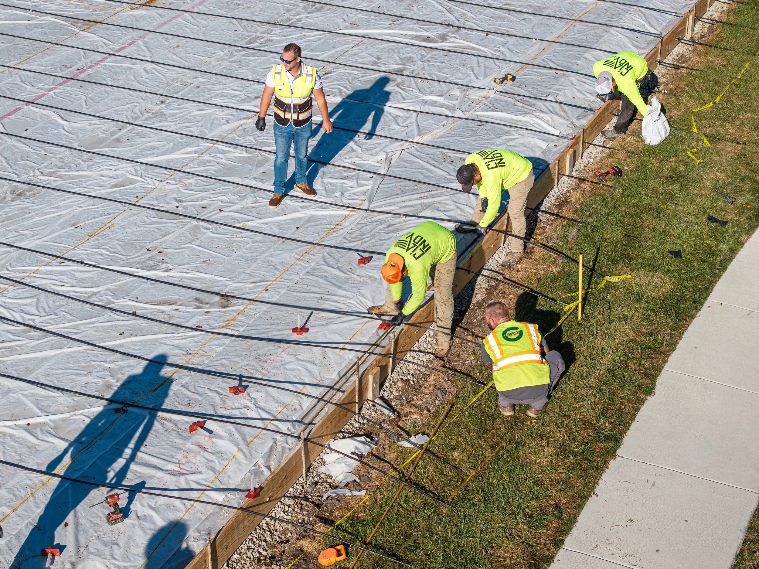 Construction workers installing or repairing a foundation with a plastic vapor barrier, several workers are bending down and securing the barrier while another worker stands observing. The workers are wearing safety vests and gloves, with some in har