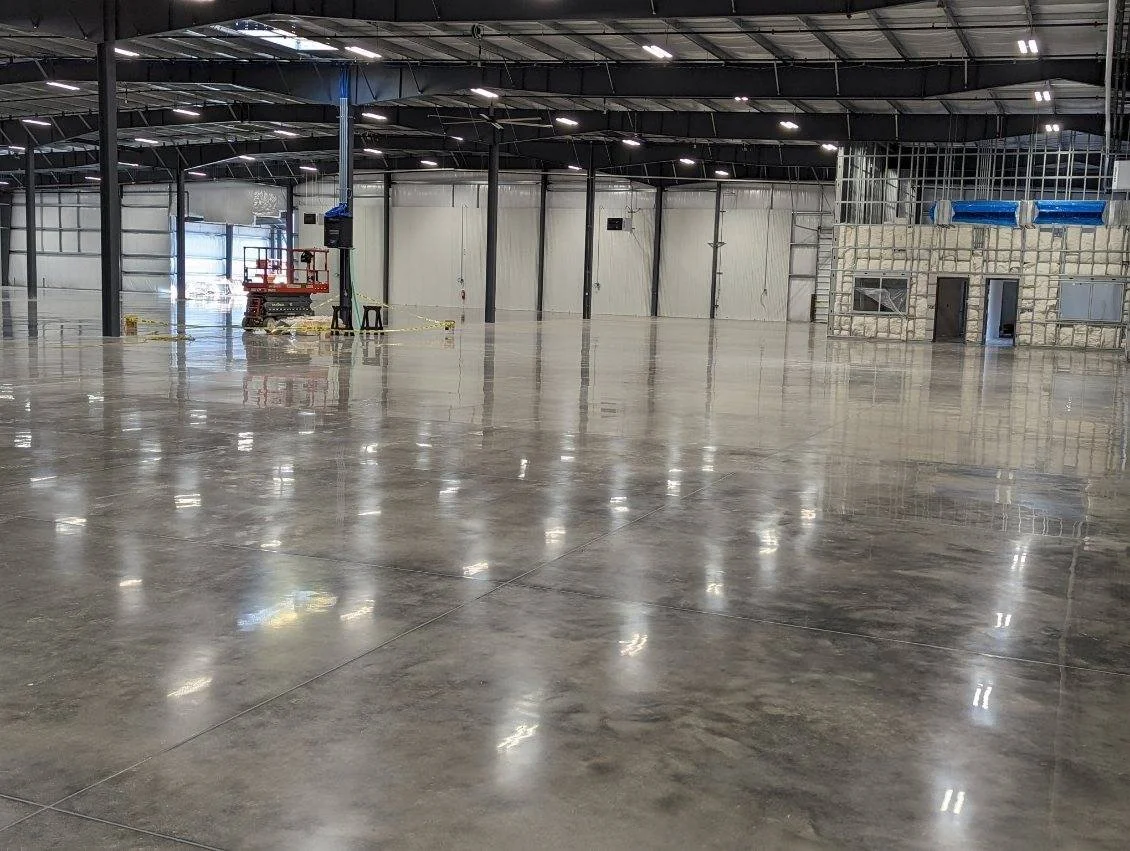 An empty warehouse with a polished concrete floor, metal walls, and a high ceiling with lights. There is a small construction area with a concrete block structure and blue ducts in the background. Equipment and yellow caution tape are also visible.