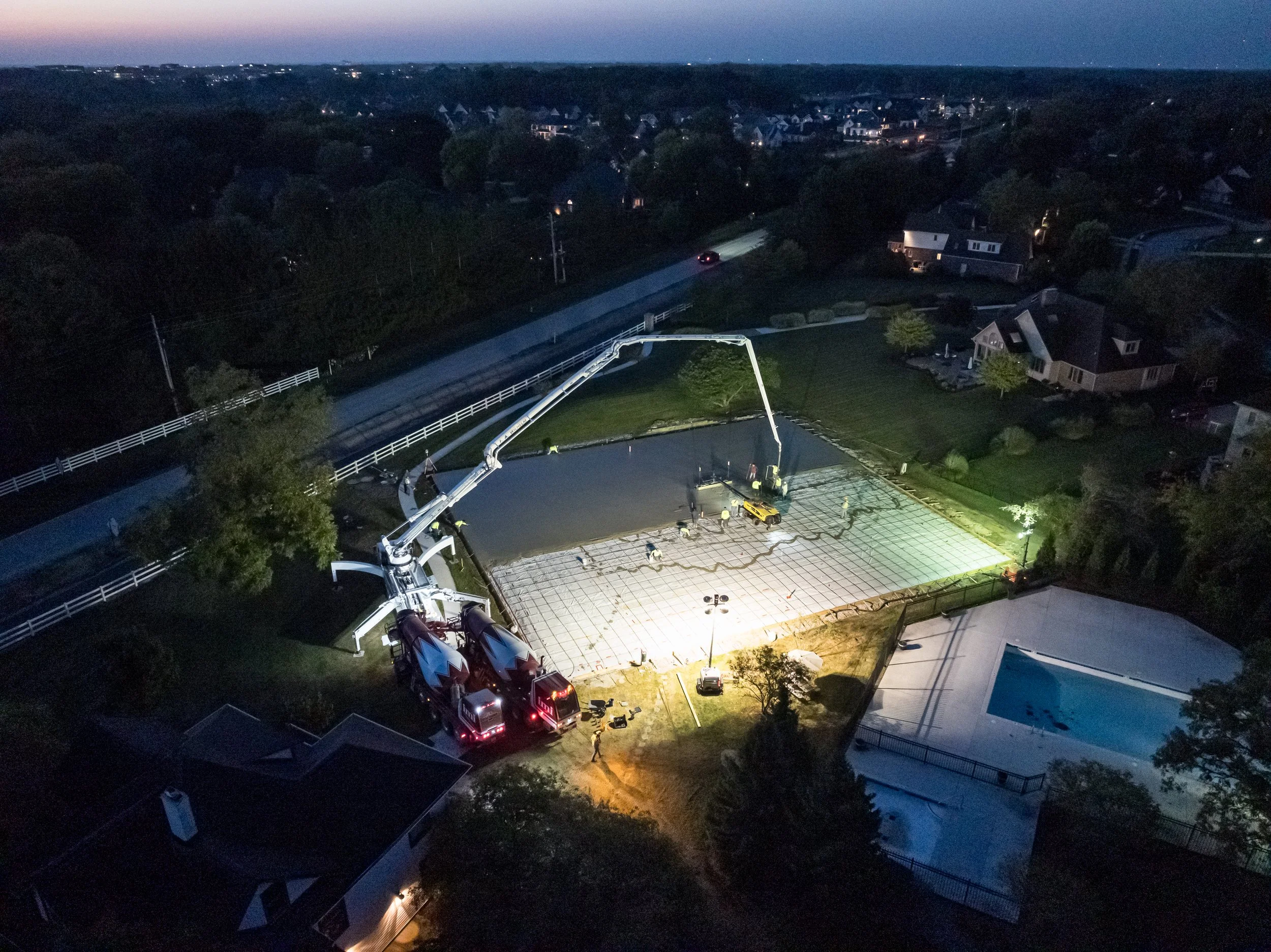 Nighttime aerial view of construction workers pouring concrete for a swimming pool with a concrete pump truck, neighboring houses, and a pool already filled with water.