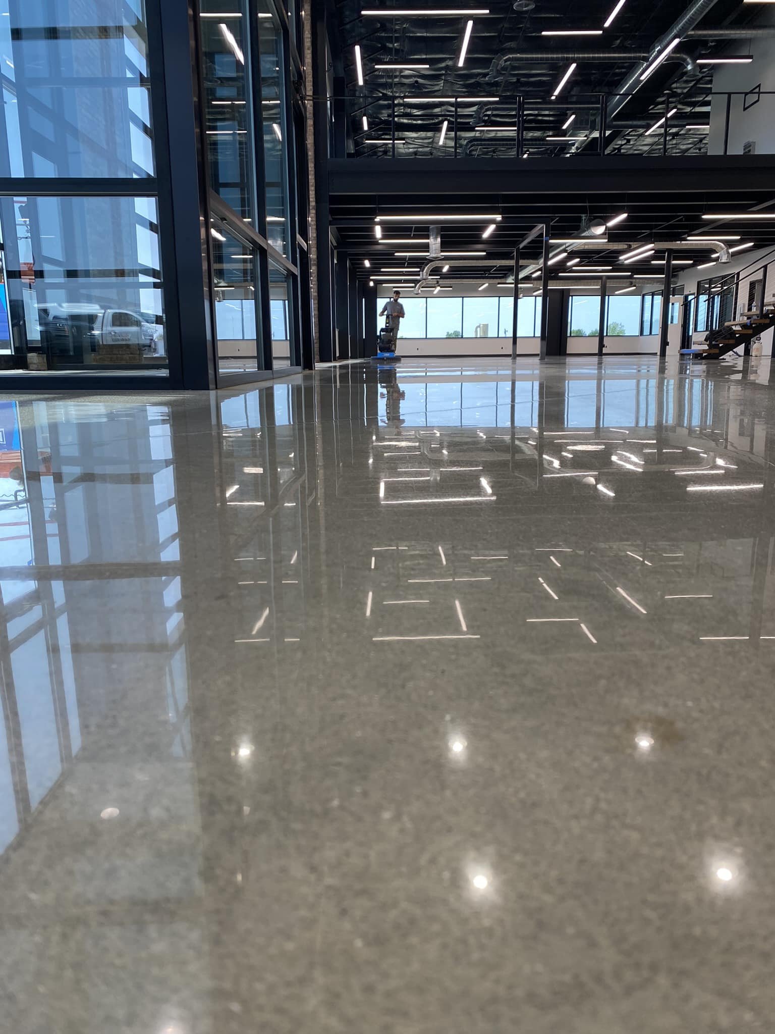 Interior of a modern, spacious building with reflective polished floors, large glass windows, and a worker cleaning the floor with a floor scrubber. The ceiling has exposed pipes and linear lighting, and there are stairs on the right side.