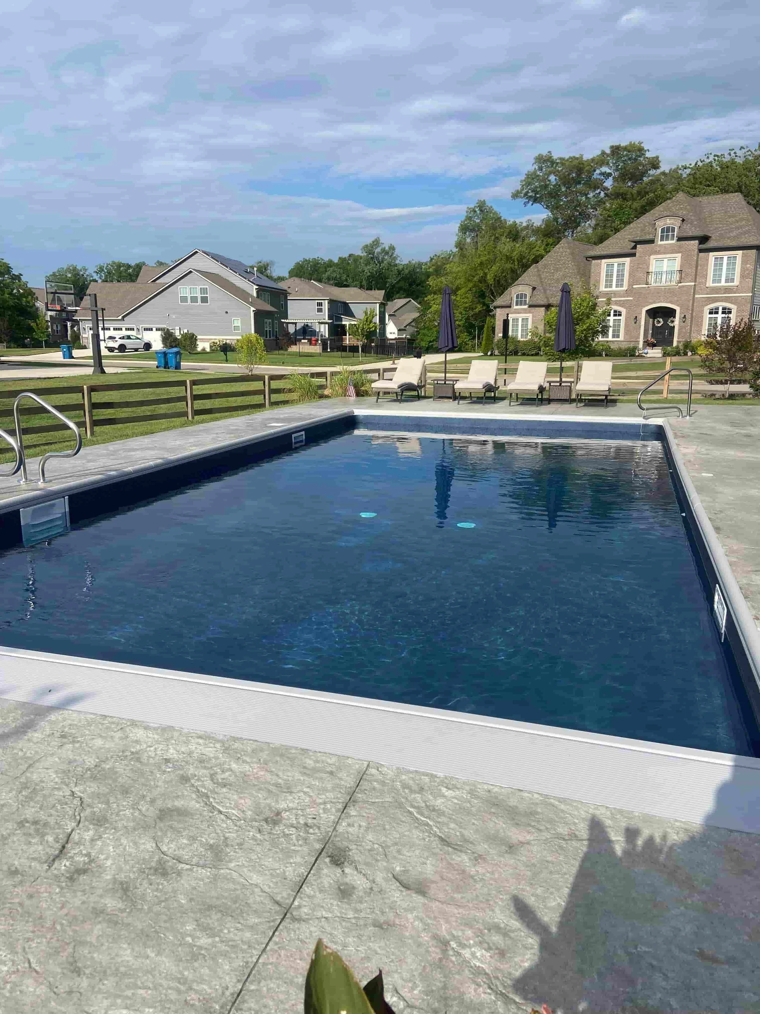 Swimming pool with lounge chairs and umbrellas in a suburban backyard under a cloudy sky.
