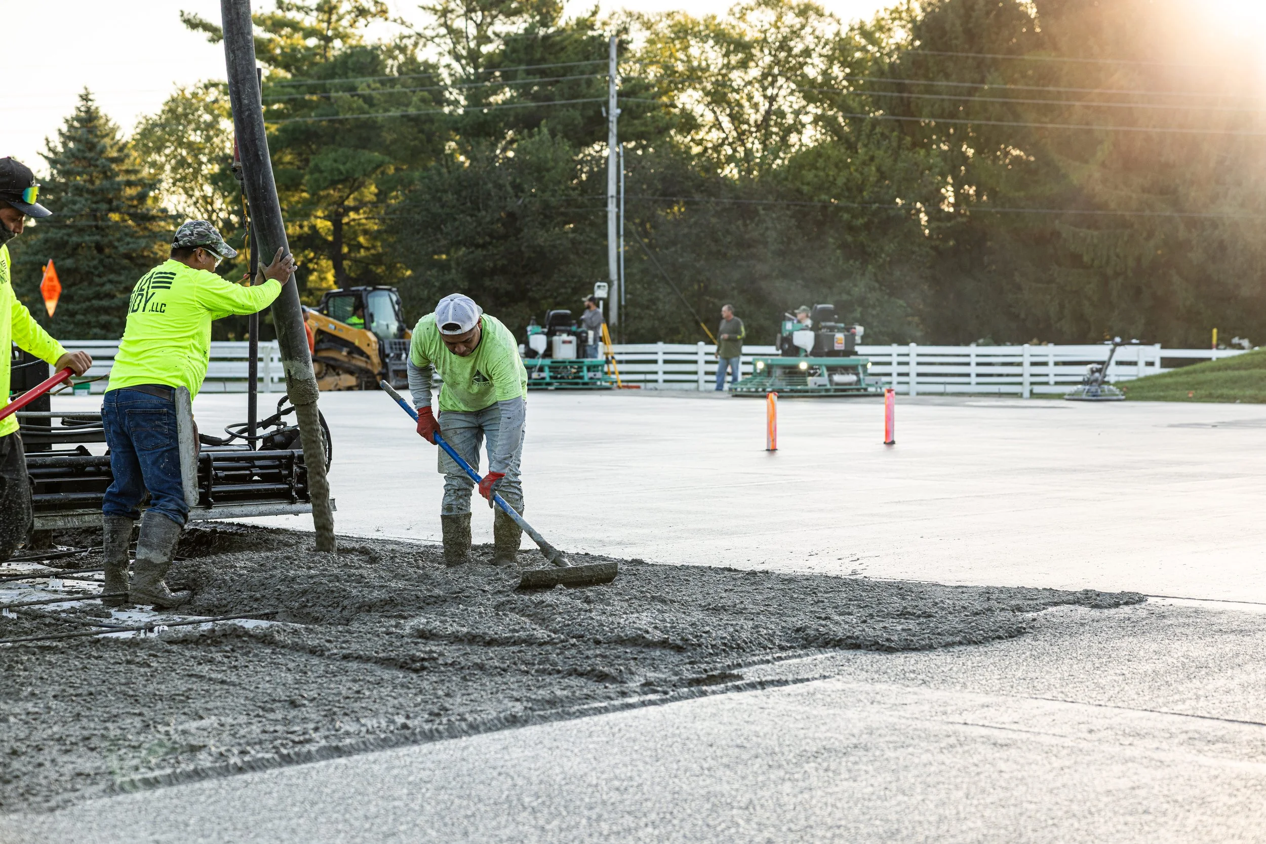 Construction workers pouring and spreading concrete on a road, with machinery and workers in the background during late afternoon.