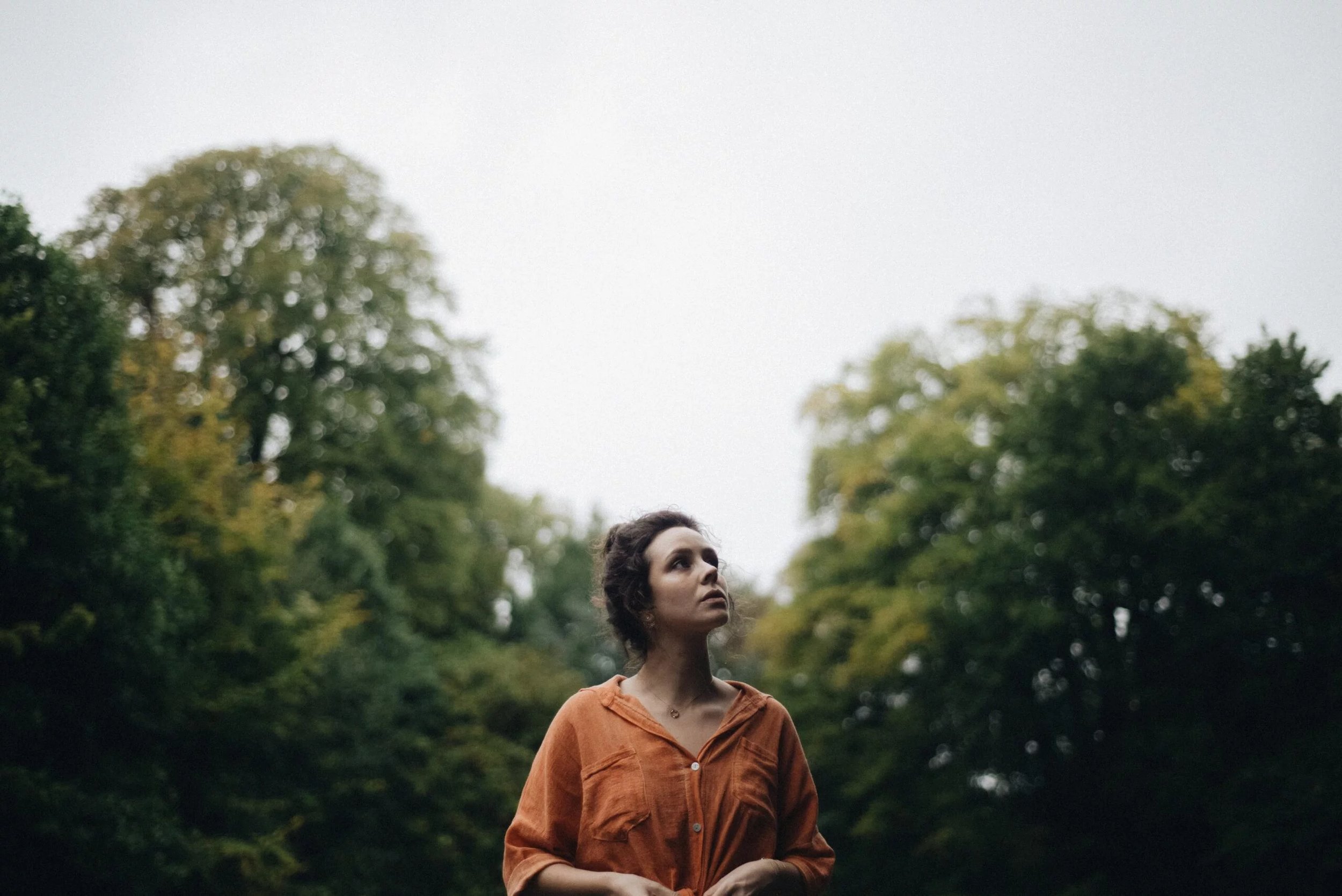 Woman in orange shirt standing in a park with trees in the background, looking up.