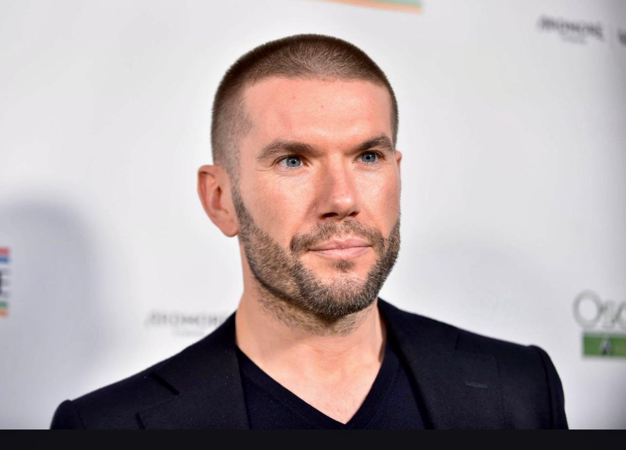 Person with a short haircut and beard, wearing a black shirt and suit, in front of a white background with logos.