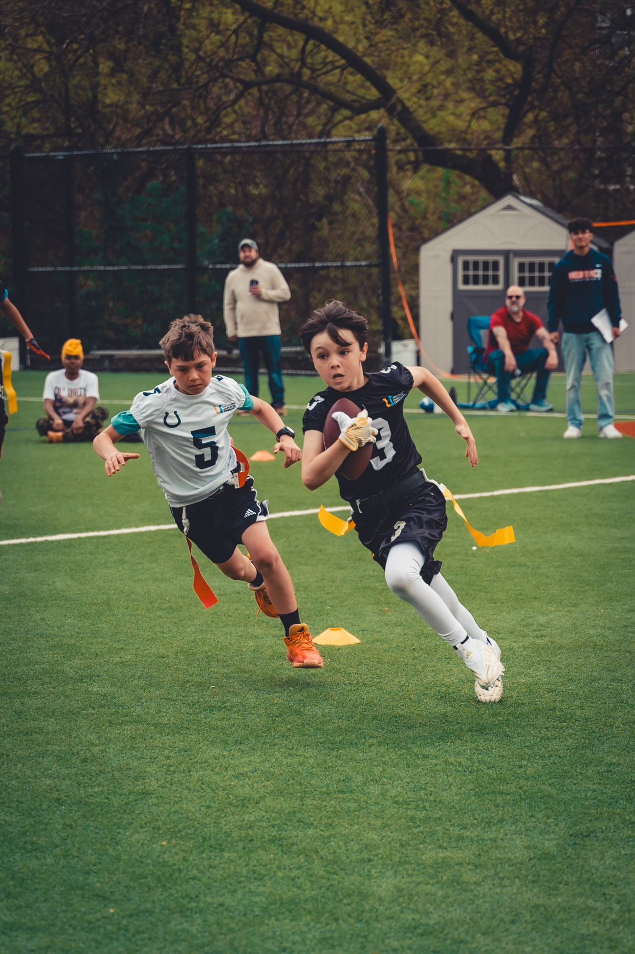 Two kids playing flag football on a grassy field, one holding the ball and wearing a black jersey, the other in a white jersey attempting to catch up. Spectators and coaches in the background.