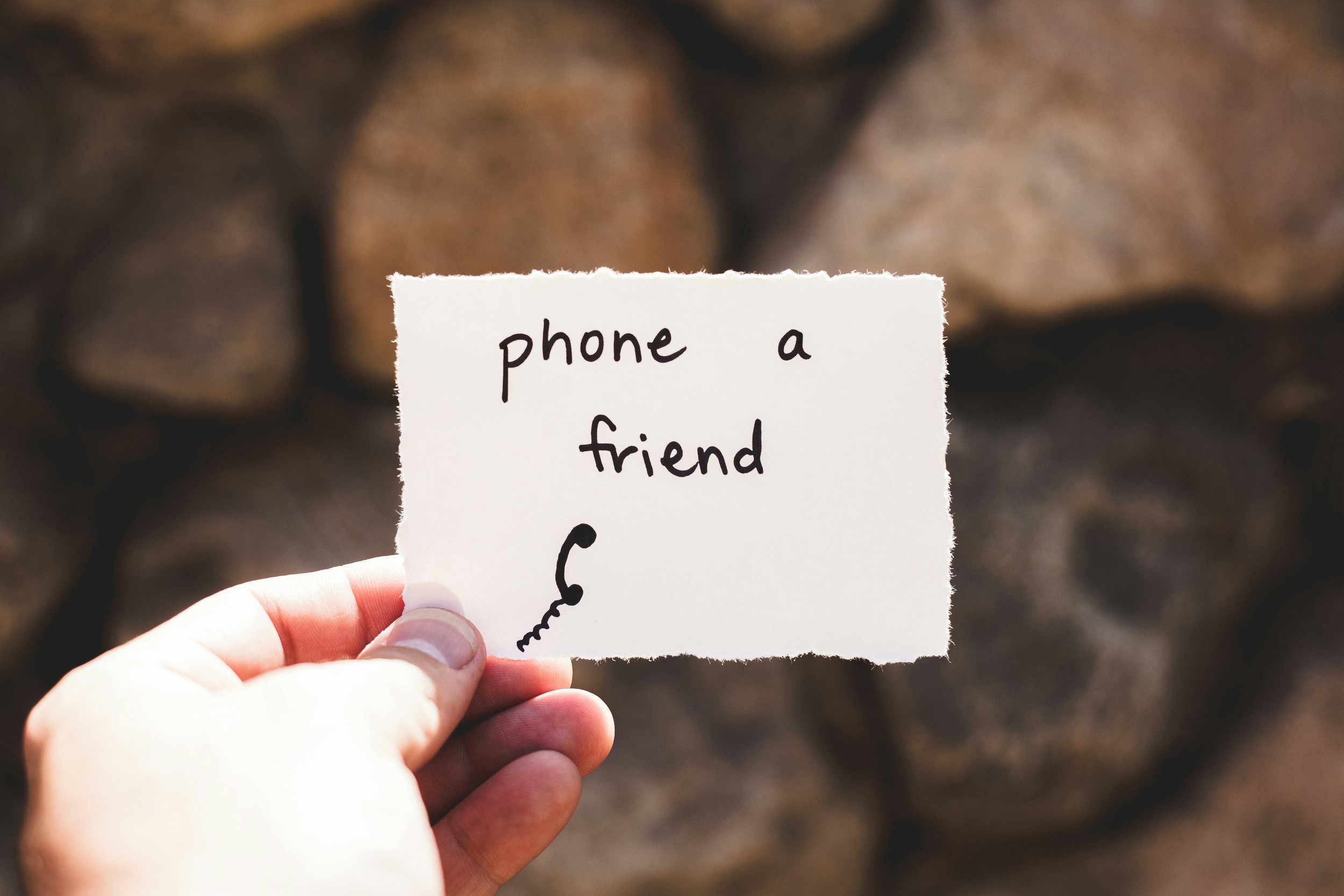 Hand holding a card with handwritten text "phone a friend" and a small phone doodle, against a blurred background.