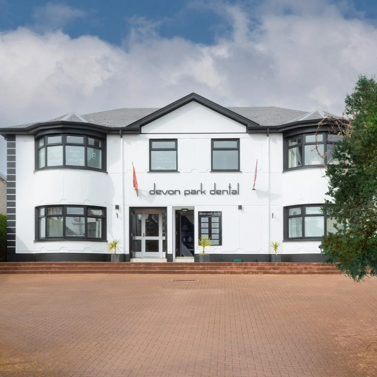 Front view of a white two-story building with black trim, labeled 'devon park dental', featuring bay windows and entrance with two potted plants outside.