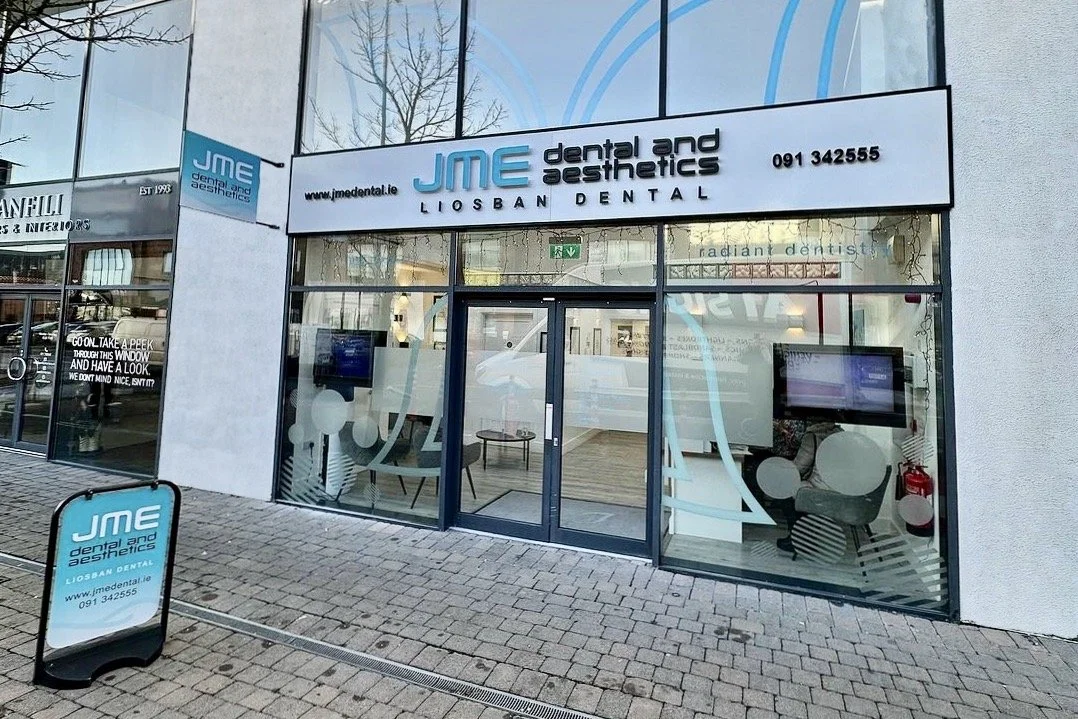 The exterior of JME Dental and Aesthetics, a dental office in Liosban, with a glass front and sign. There is a signboard outside with the clinic's name and contact information.