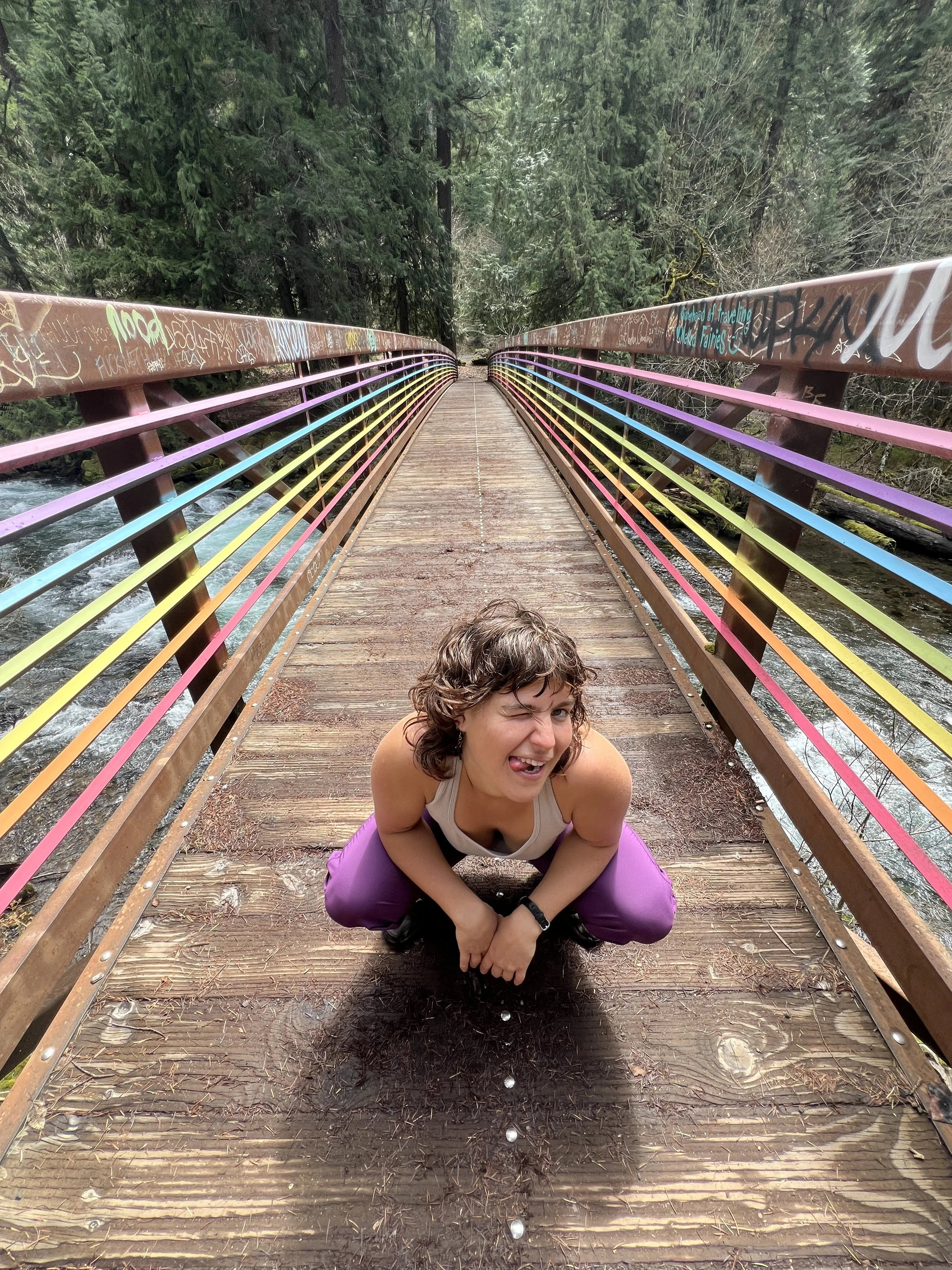 A person squatting on a colorful bridge with graffiti, surrounded by trees, making a playful expression.