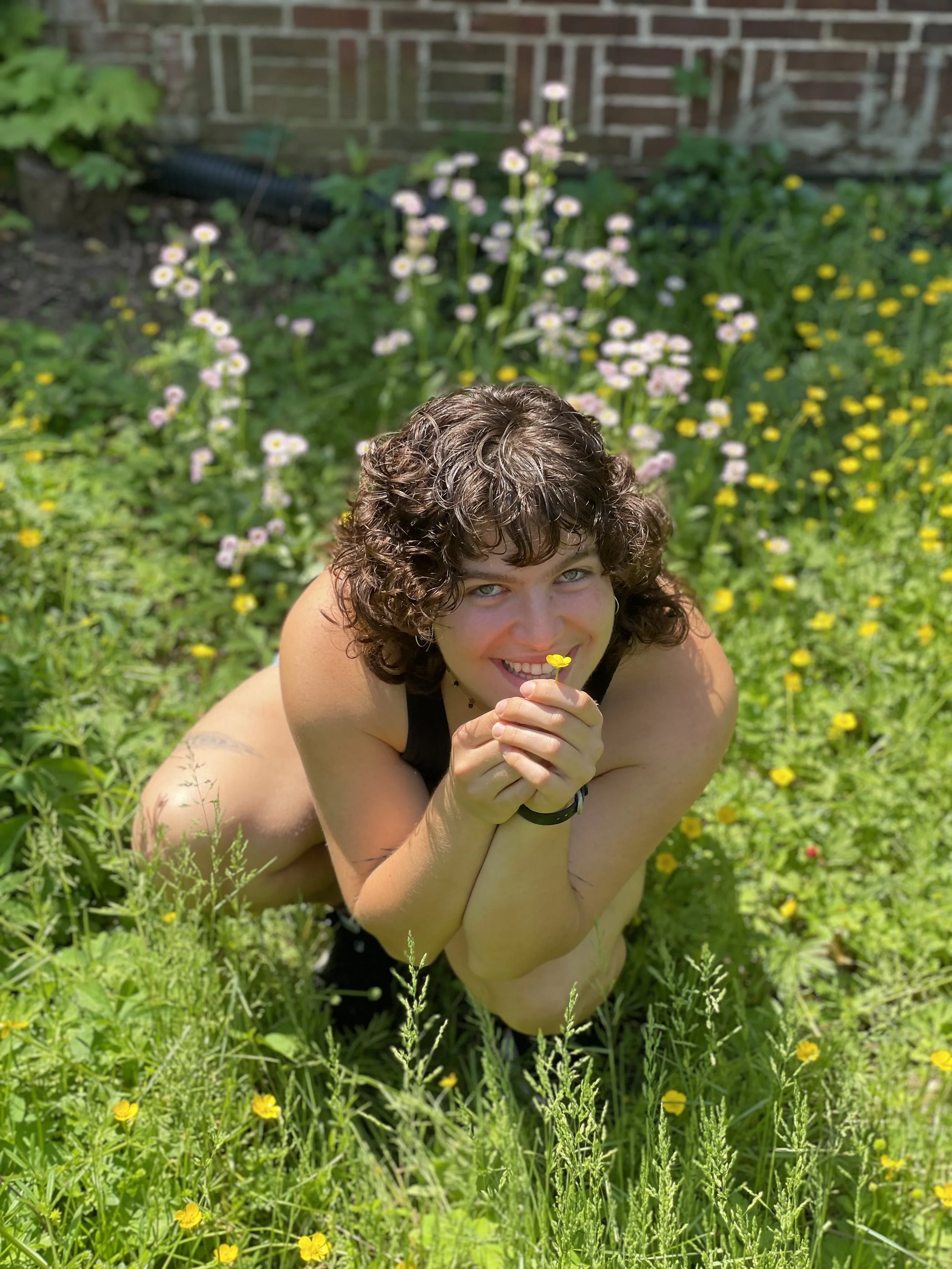 Person crouching in a garden holding a yellow flower, surrounded by green plants and multiple pink wildflowers.
