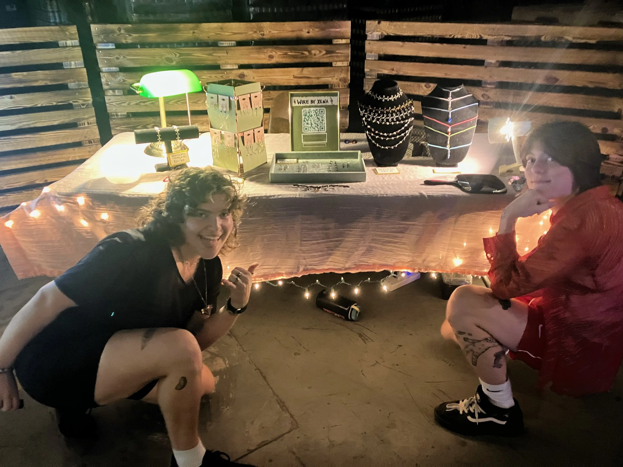 Two people kneeling in front of a table decorated with string lights, displaying jewelry and cards. The setting suggests a cozy, informal market stall or exhibition.