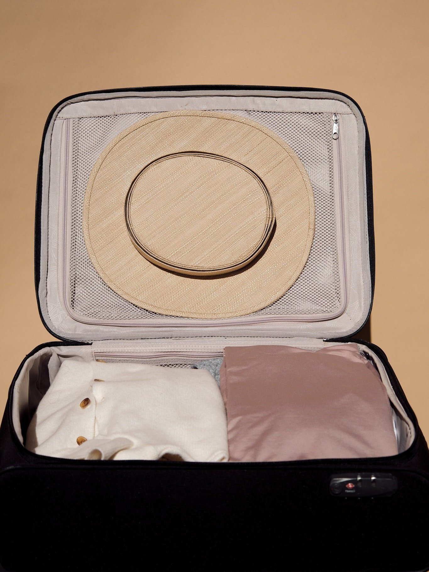 Open suitcase with neatly packed beige hat, white shirt, and pink fabric against a beige background.