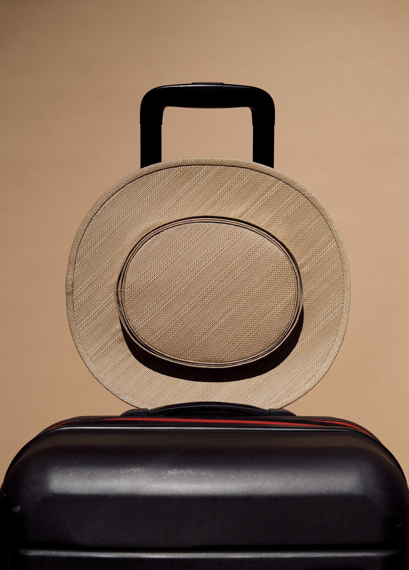 Beige straw hat on top of black suitcase against light brown background.