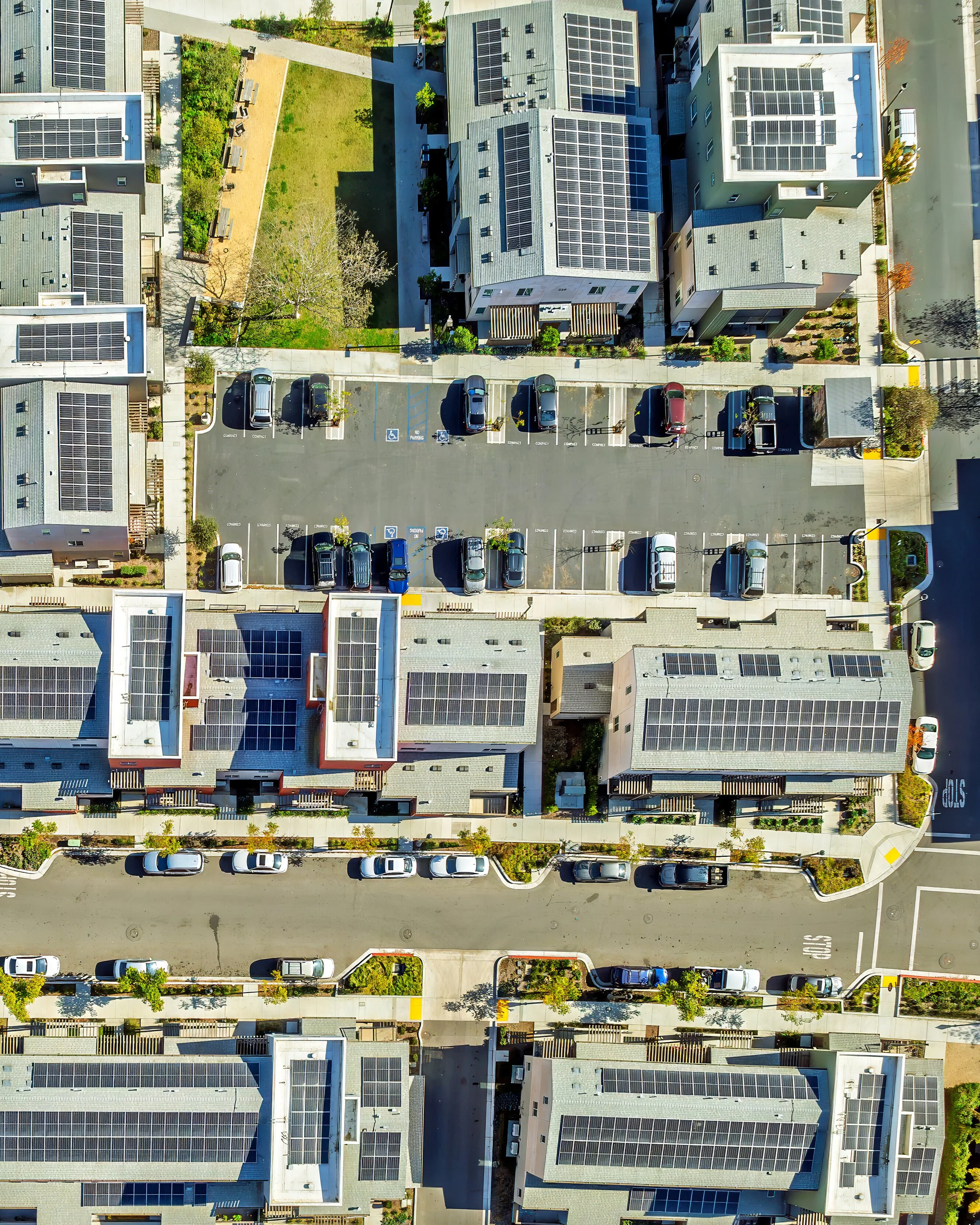 Birds-eye view of a multi-family housing unit with solar on 10 plus different buildings, plus a parking lot with cars.