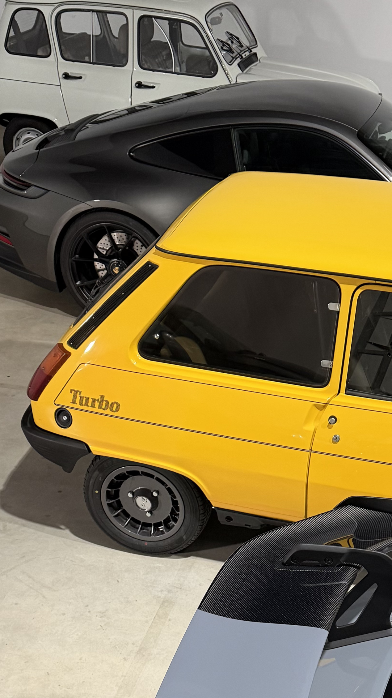 Drei verschiedene klassische Autos im Showroom, darunter ein gelber Renault mit dem Aufdruck 'Turbo', ein grauer Porsche und ein weißer Citroën.