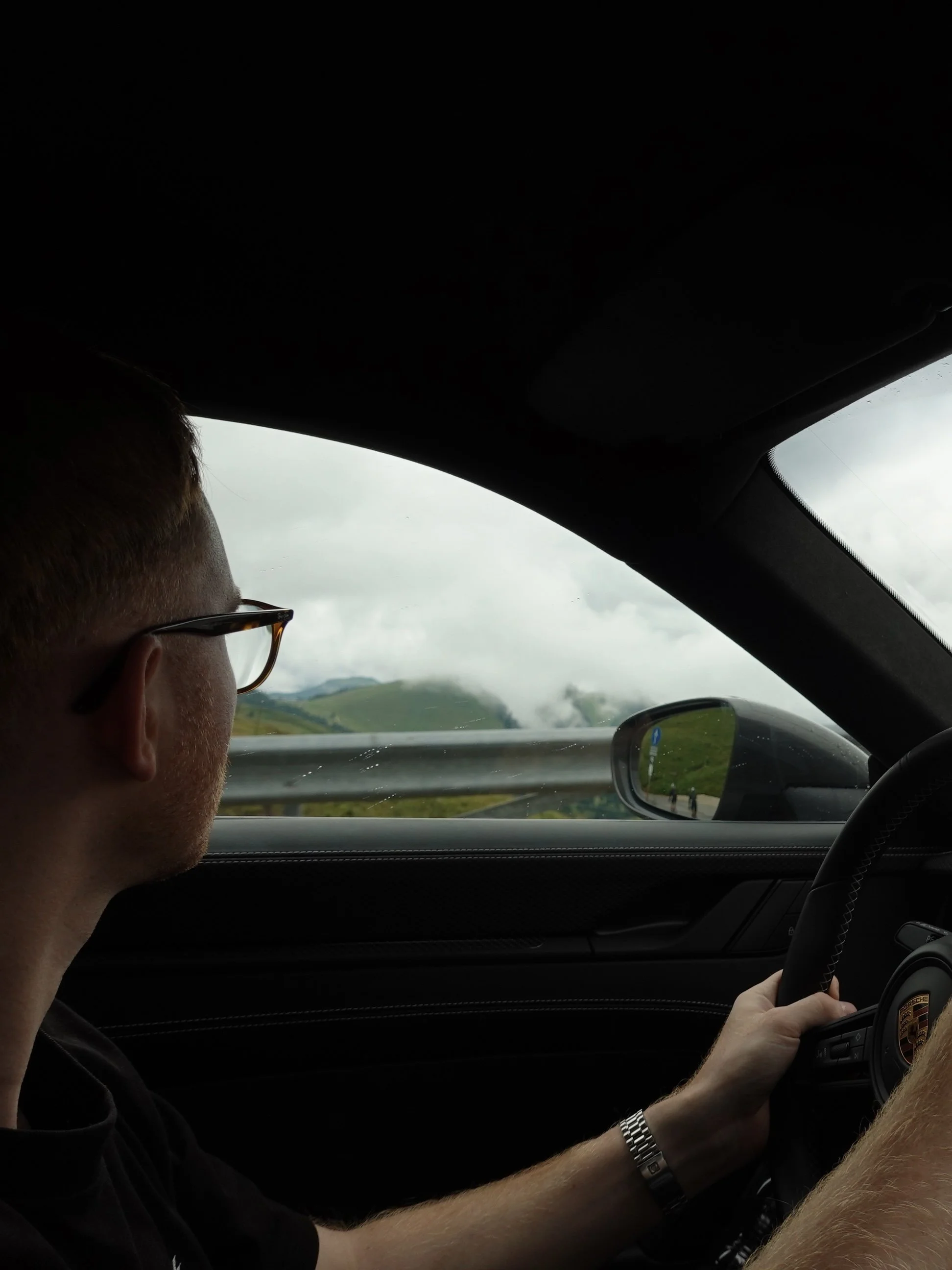 Der Fahrer eines Porsche-Fahrzeugs fährt auf einer Landstraße durch eine ländliche Landschaft mit grünen Hügeln und bewölktem Himmel.