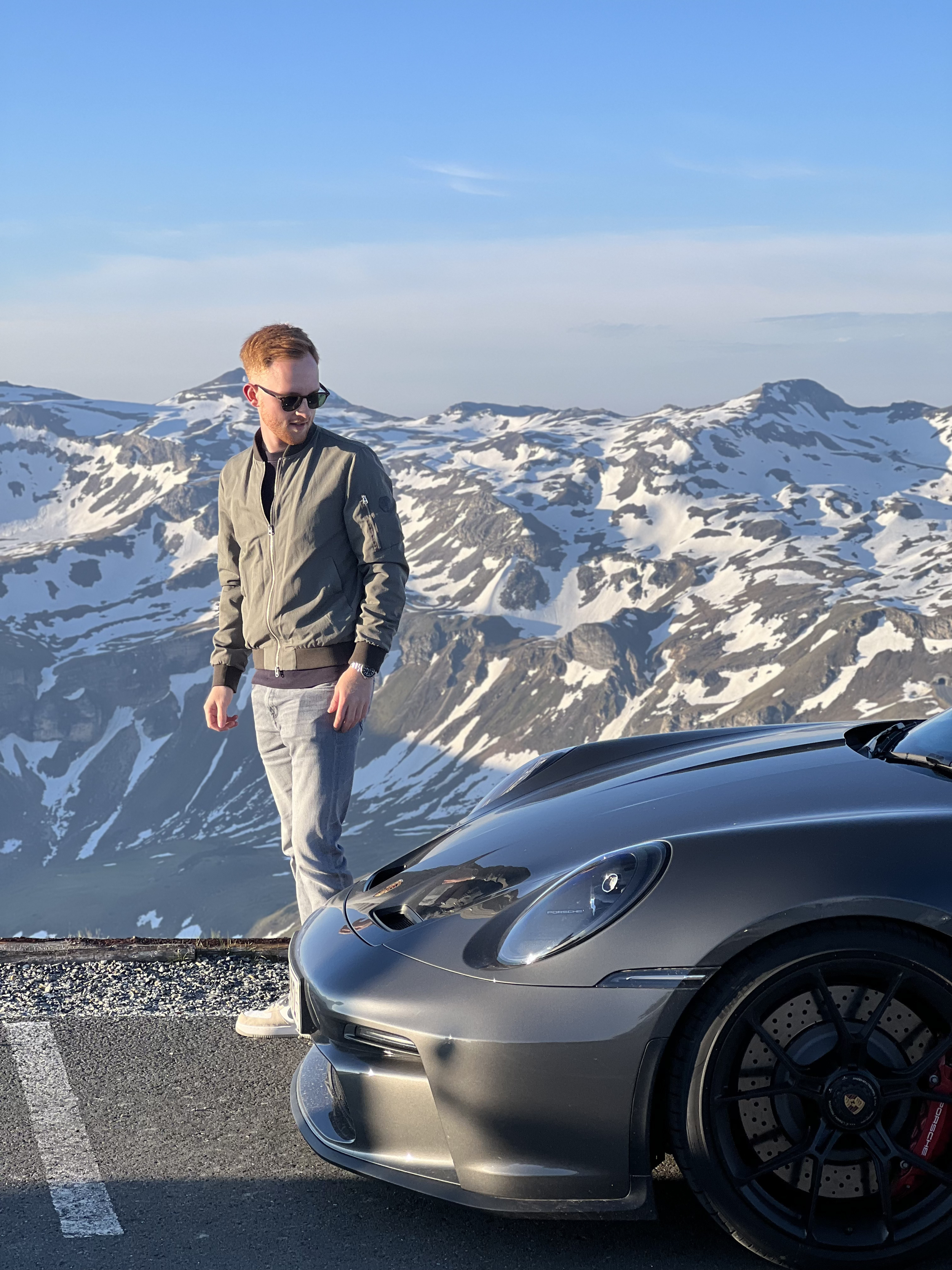 Ein junger Mann mit Sonnenbrille steht neben einem silbernen Porsche in einer bergigen Schneelandschaft bei Aufgang.