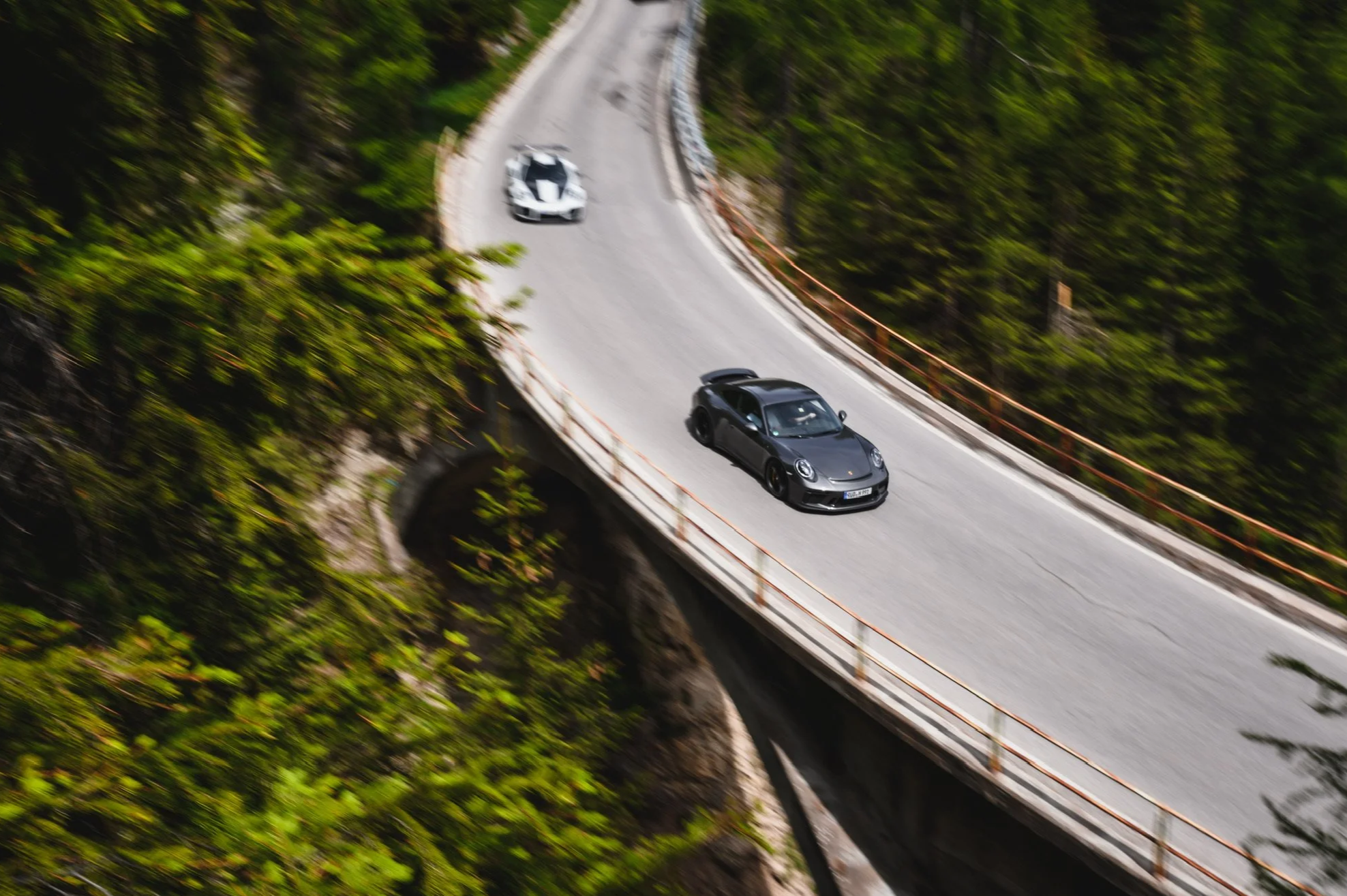 Zwei Sportwagen fahren auf einer kurvigen Bergstraße zwischen Bäumen.