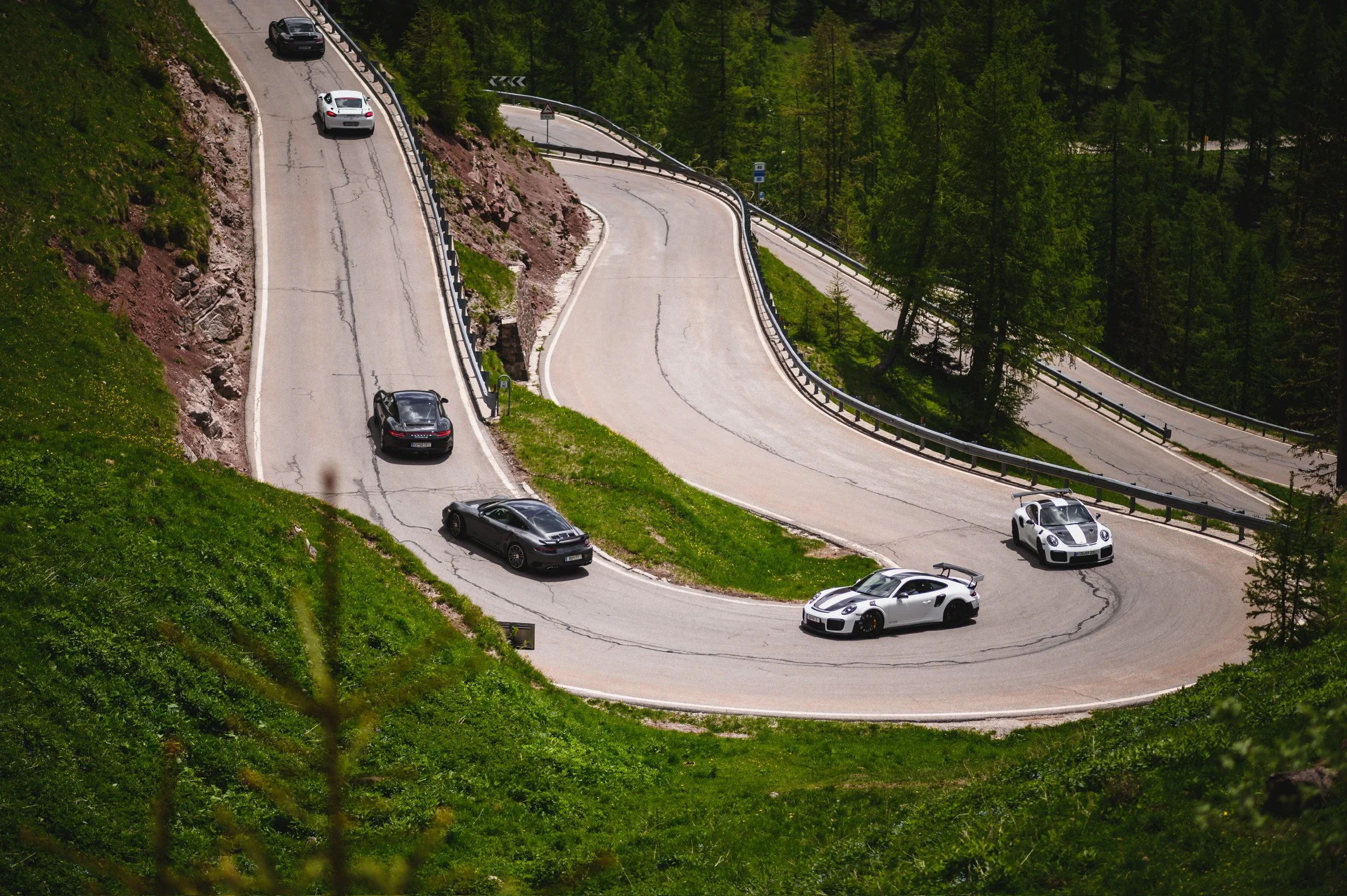 Scharfe Kurven auf einer talseitigen Bergstraße, mehrere Autos, üppige grüne Wäldern im Hintergrund.