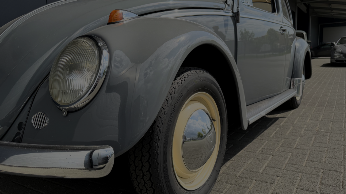 Nahaufnahme eines klassischen grauen VW Käfer, Fokus auf Vorderseite und Reifen, im Freien geparkt auf einer gepflasterten Fläche.