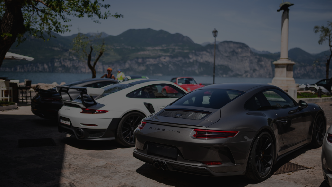 Drei Sportwagen sind in einer Küstenstadt parkend, mit Wasser und Bergen im Hintergrund.