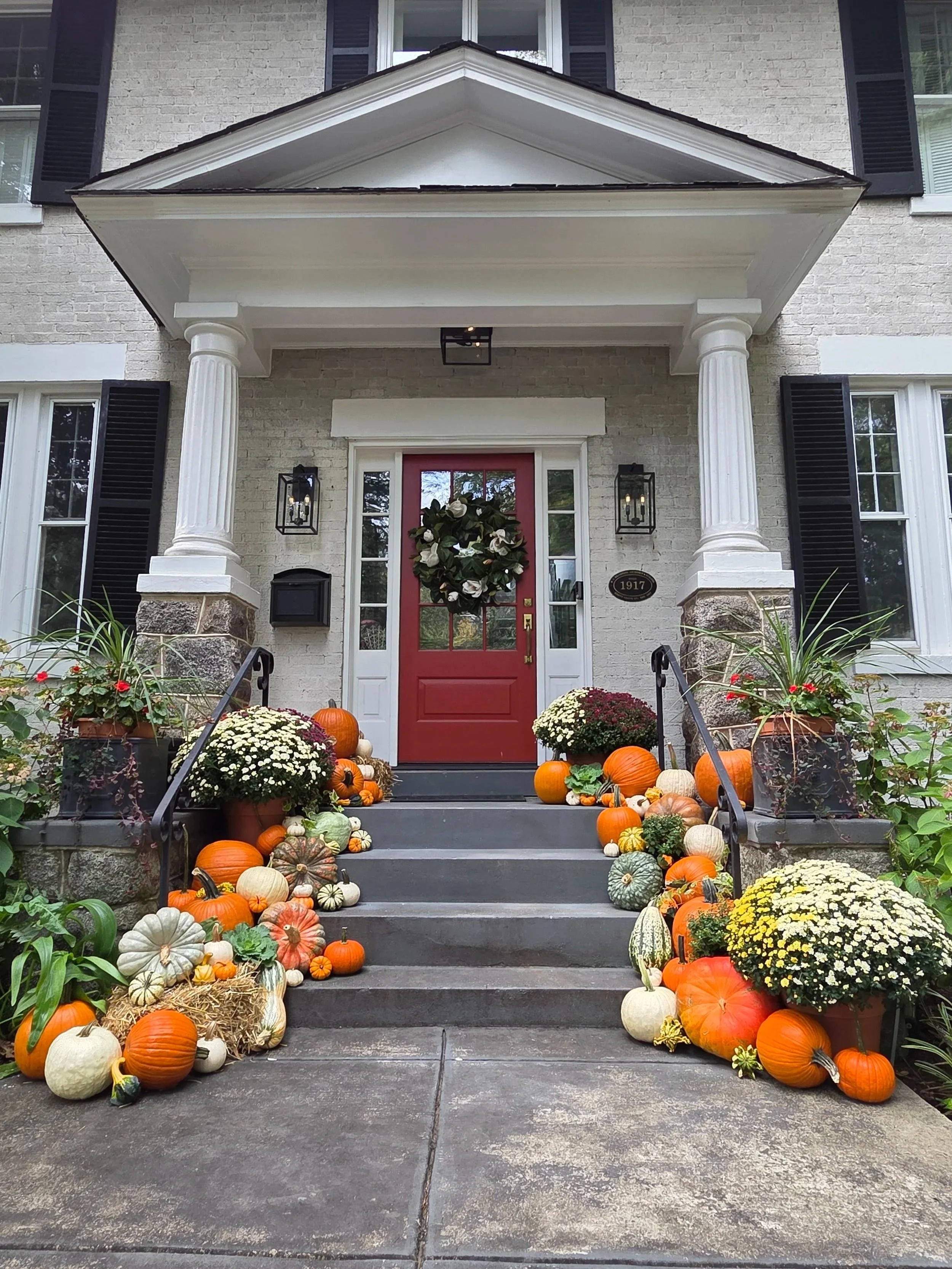 Front steps of a house decorated with pumpkins and flowers for fall or Halloween, including chrysanthemums and marigolds, with a wreath on the red front door.