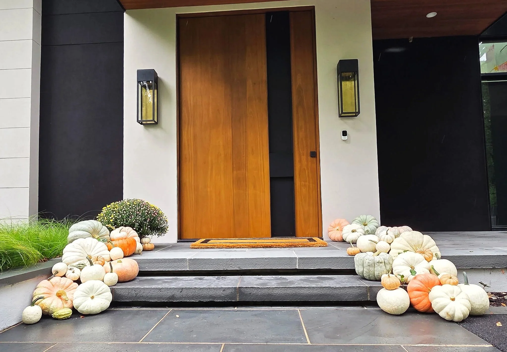 Front porch with a wooden door flanked by two lantern-style wall lights, a welcome mat, pumpkin decorations on steps and plants on either side, and a dark tile walkway.