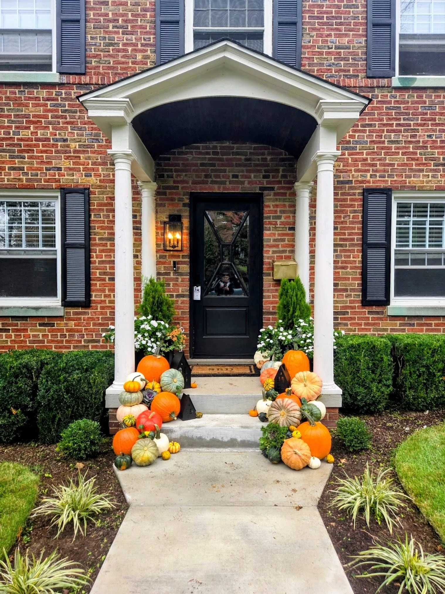 Front porch decorated with pumpkins, gourds, and fall plants for autumn or Halloween, with a brick house and black front door.