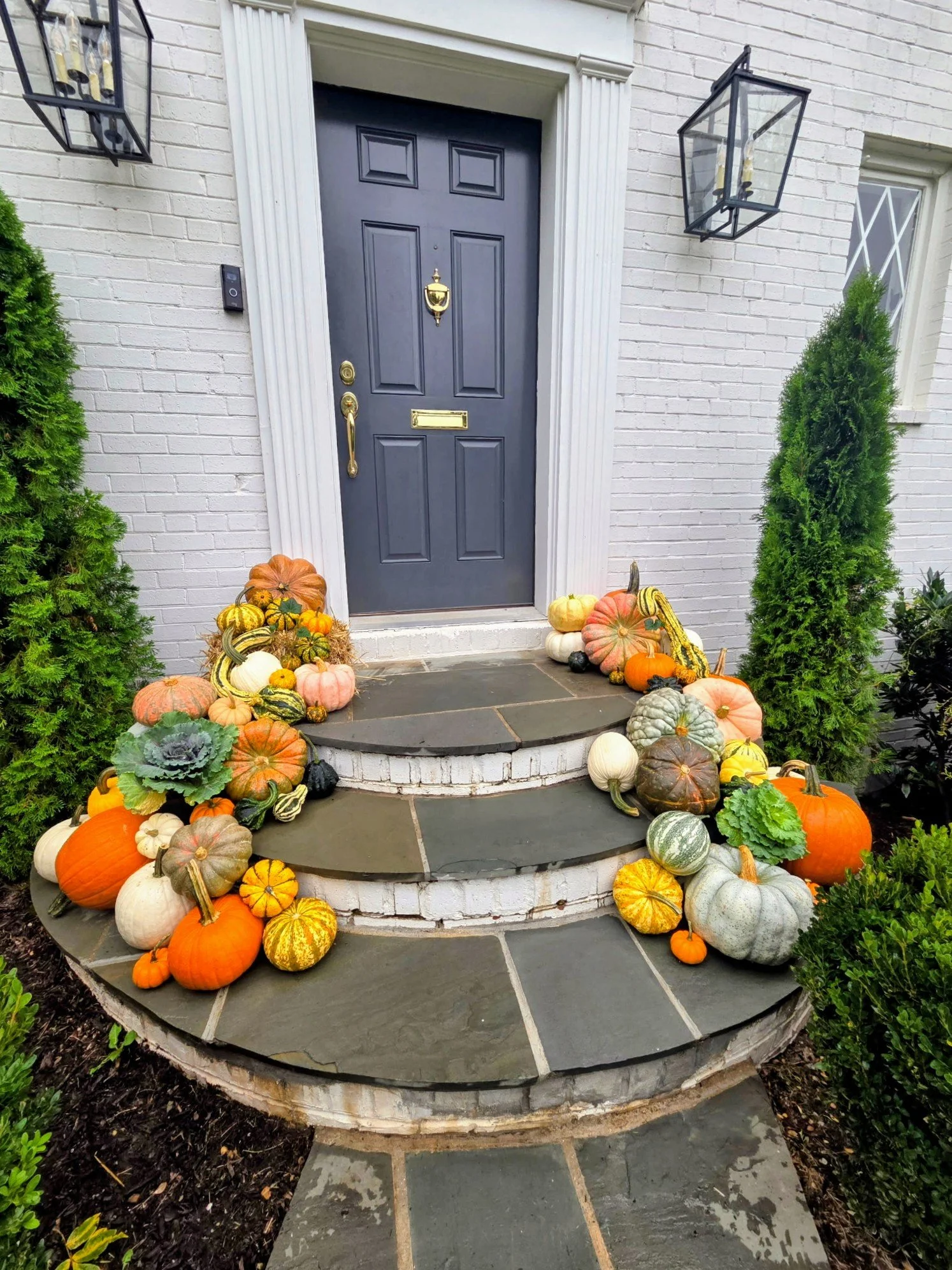 A black front door with a gold mail slot, door knocker, and handle, surrounded by white brick walls. The door is flanked by two black outdoor lanterns. The steps leading to the door are decorated with a variety of pumpkins and gourds, and there are g