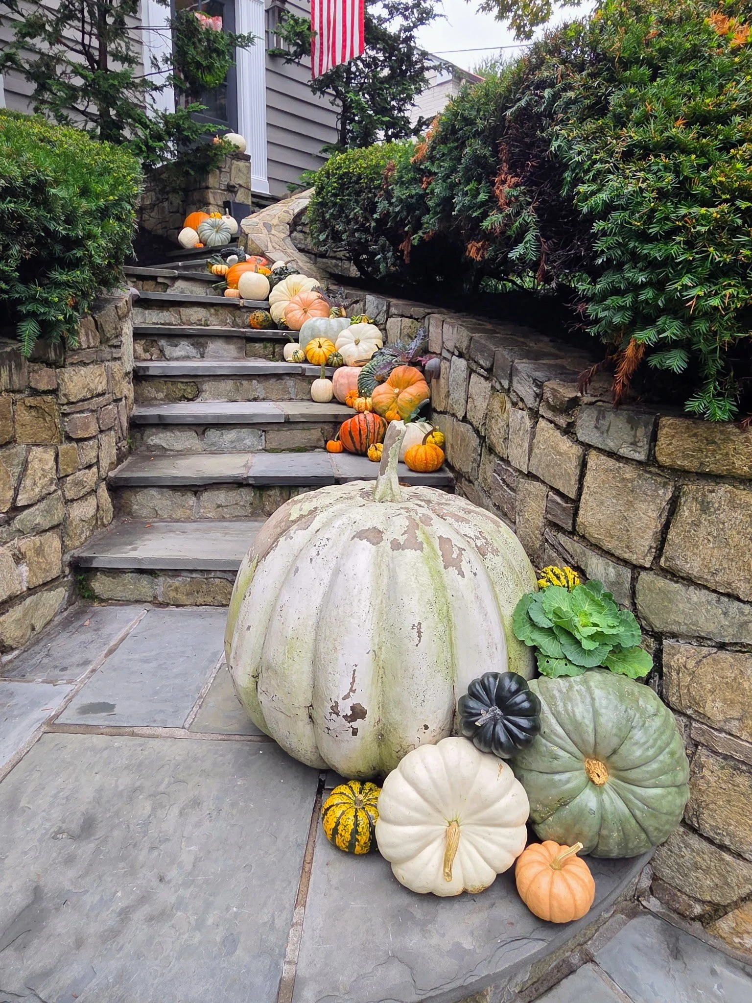 Various pumpkins and gourds arranged on stone steps and a large pumpkin at the bottom of the stairs, with green bushes on either side, and a house with gray siding and an American flag in the background.