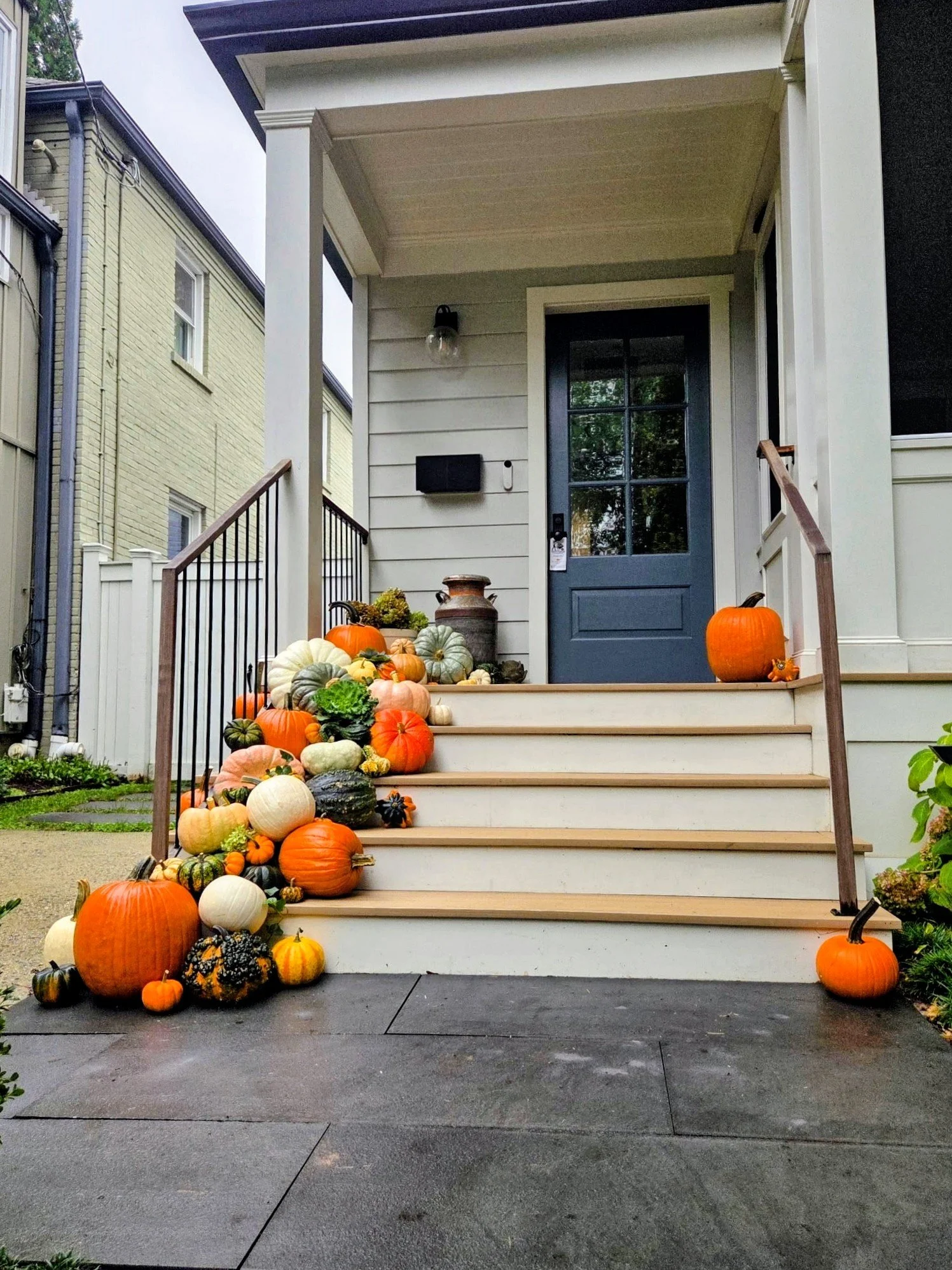 Front porch with stairs decorated with various pumpkins and gourds for fall or Halloween. The house has white siding, a dark blue door, and a small black mailbox. There are potted plants and a decorative urn on the porch.