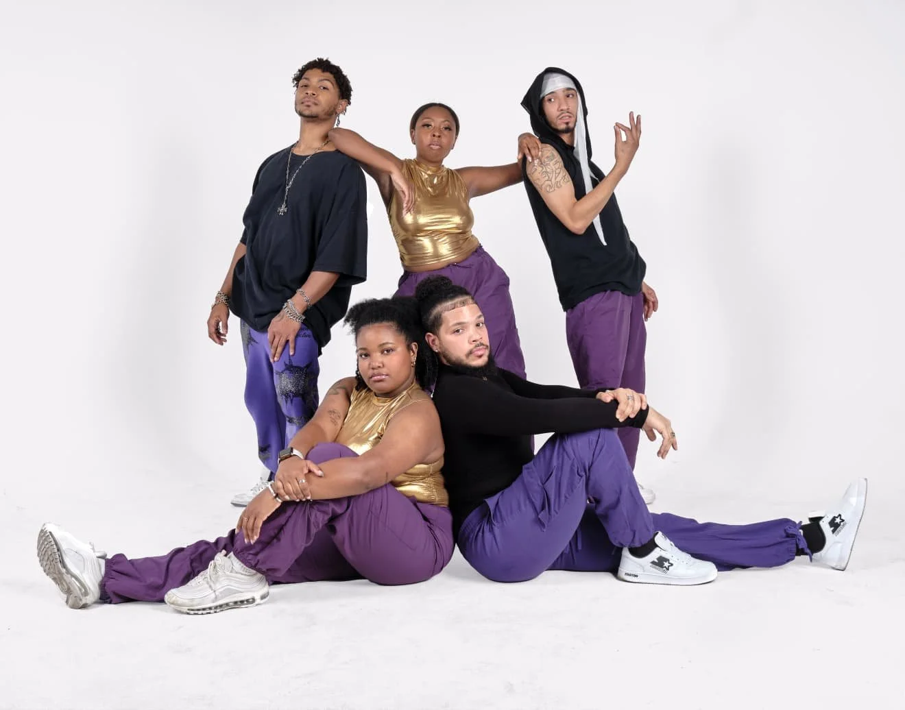 Five people posing together in casual streetwear, with a clean white background. Two individuals are seated on the floor, while three are standing behind them, some with their arms around each other.