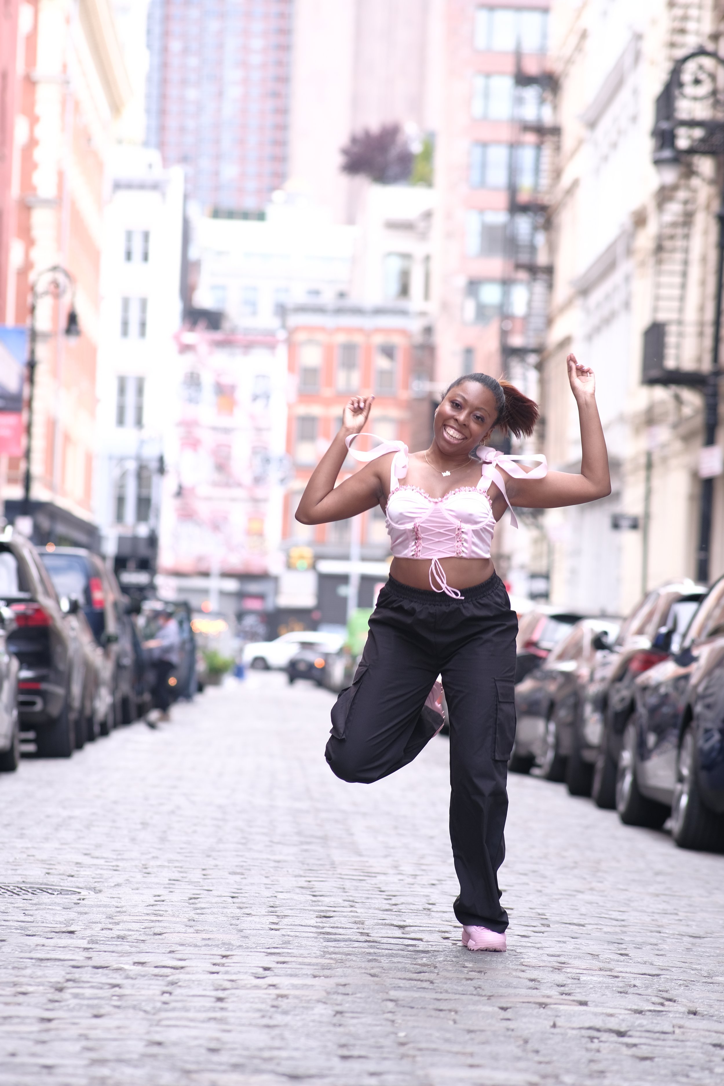 Young woman smiling and jumping in the middle of an urban cobblestone street surrounded by parked cars, with tall buildings in the background.