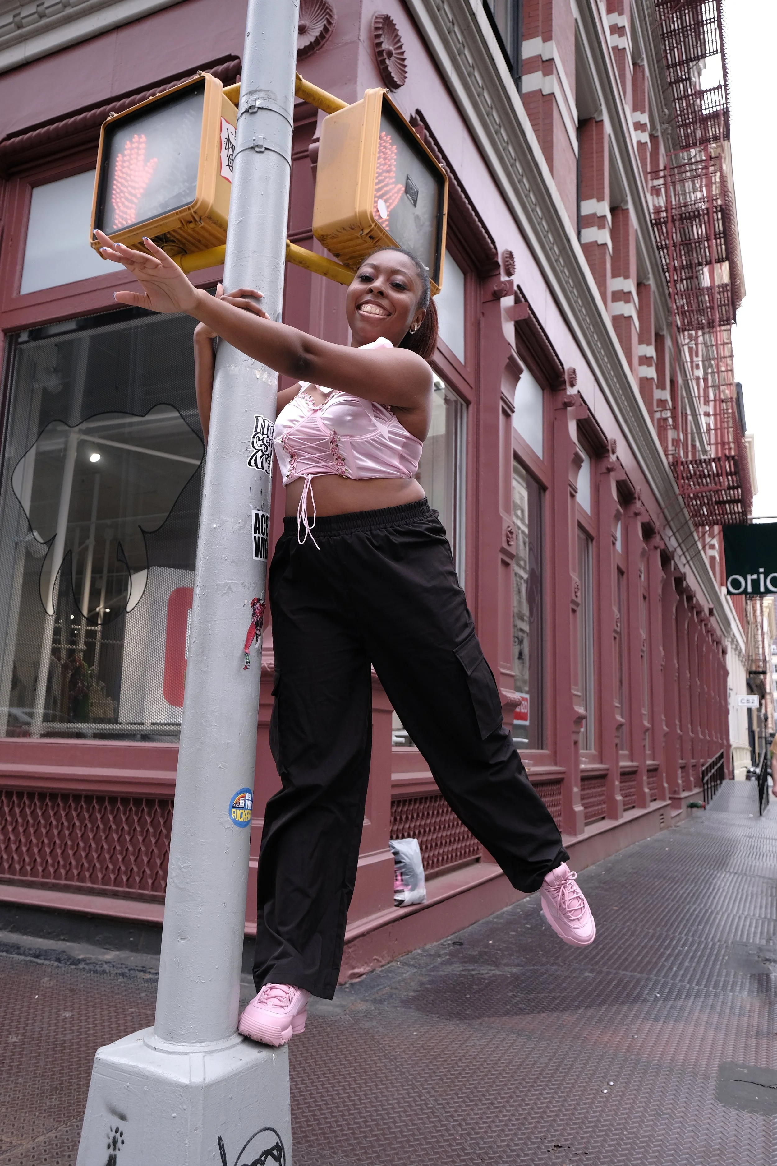 A smiling woman in pink sneakers, black pants, and a pink top, hugging a streetlight pole on a city sidewalk, with a red brick building in the background.