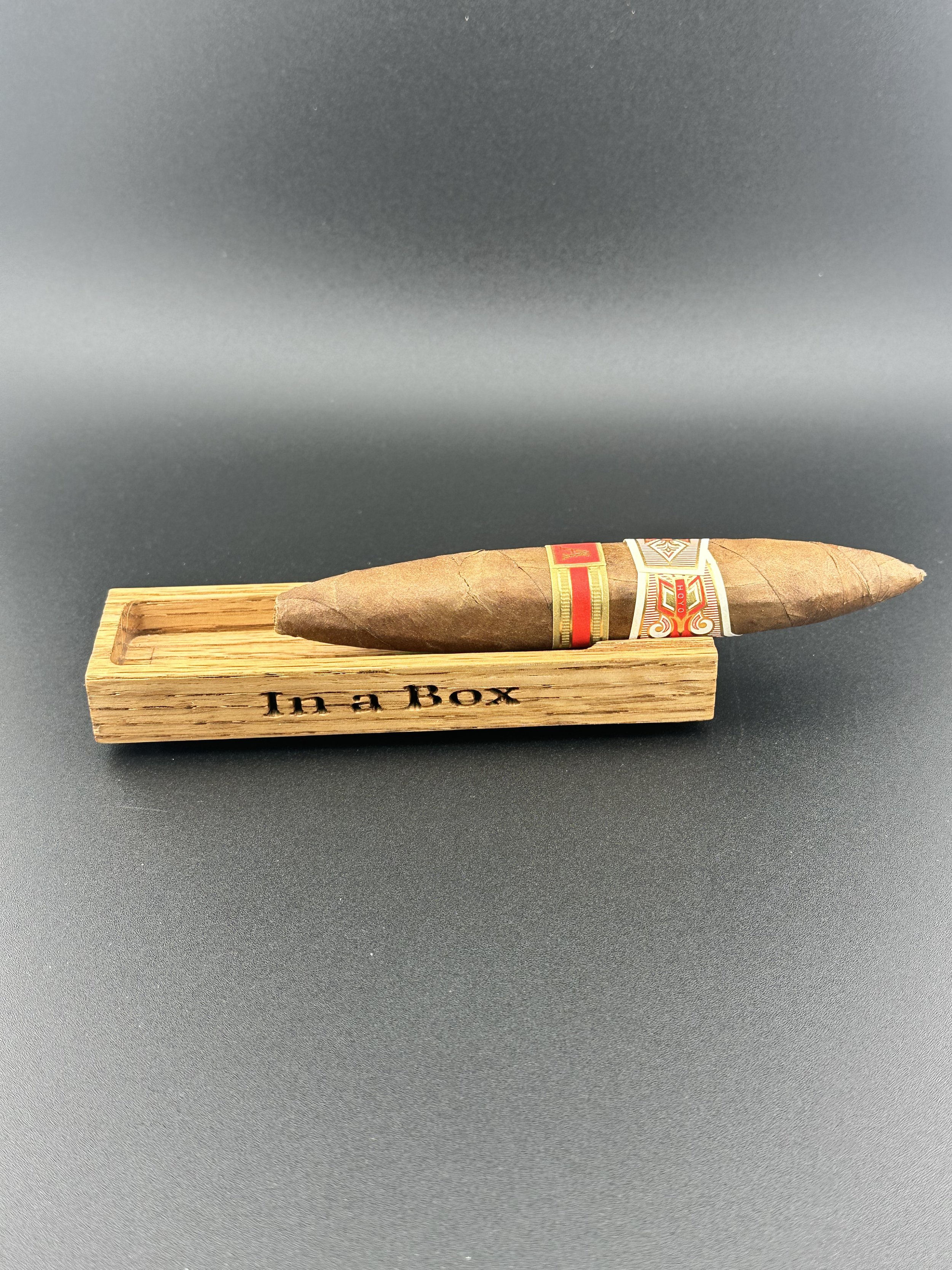 Cigar resting on a wooden box with the engraved words 'In a Box' against a plain dark background.