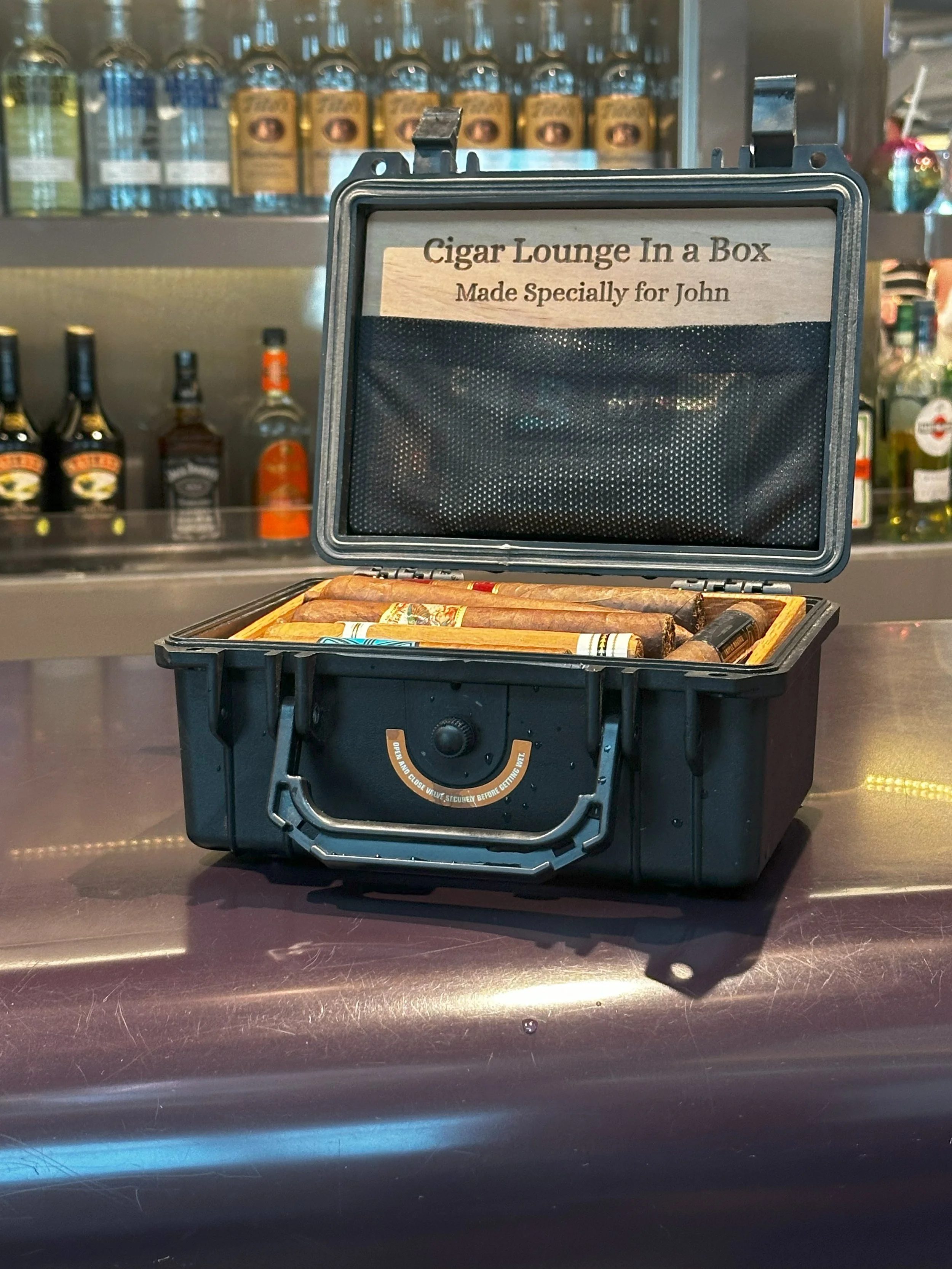 A black portable cigar case with cigars inside, labeled 'Cigar Lounge In a Box Made Especially for John,' sitting on a bar counter with bottles of alcohol in the background.