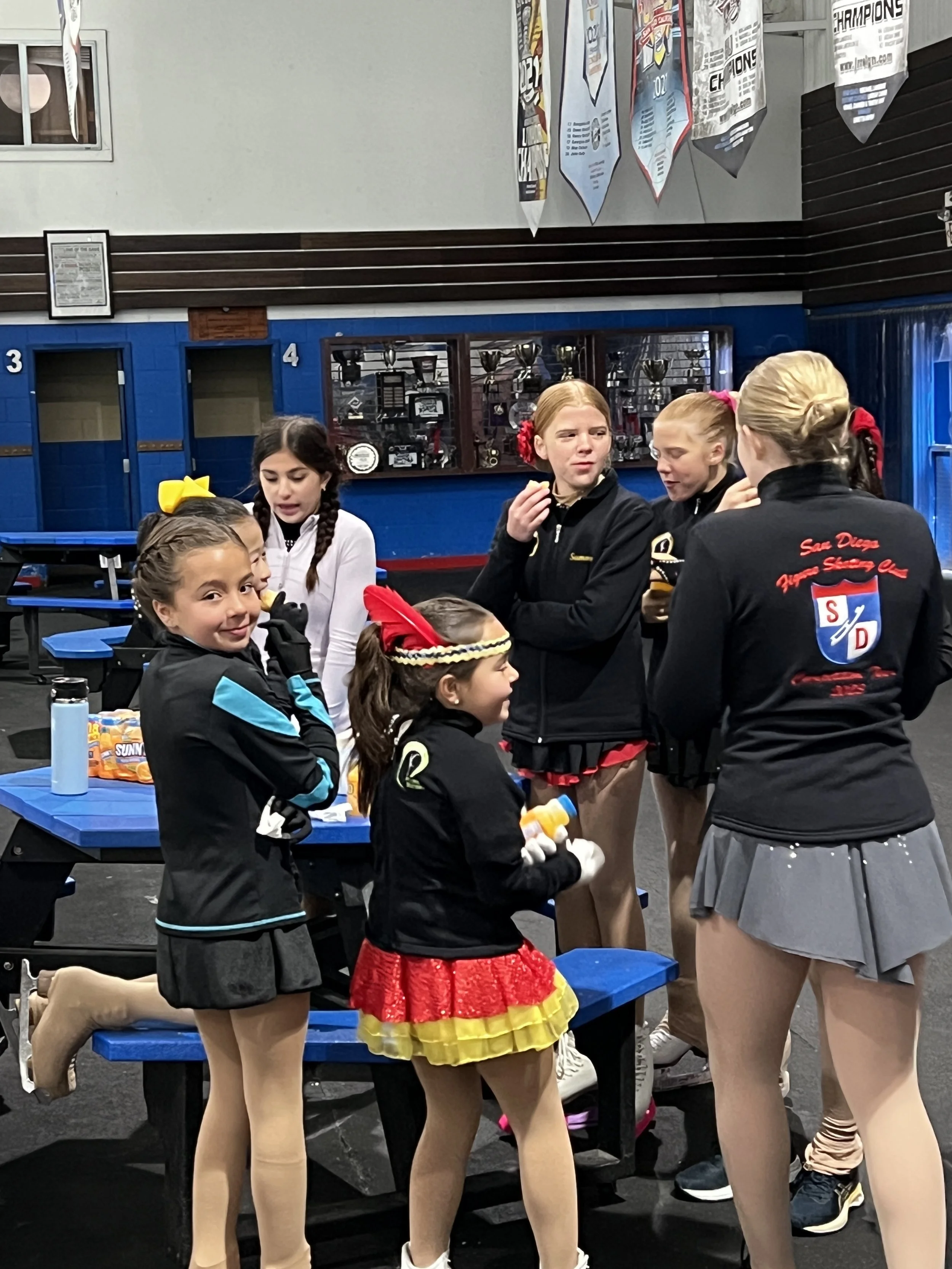 A group of young girls in cheerleader outfits gathered around in a gym or training center, some holding water bottles, with blue benches and a display of trophies in the background.