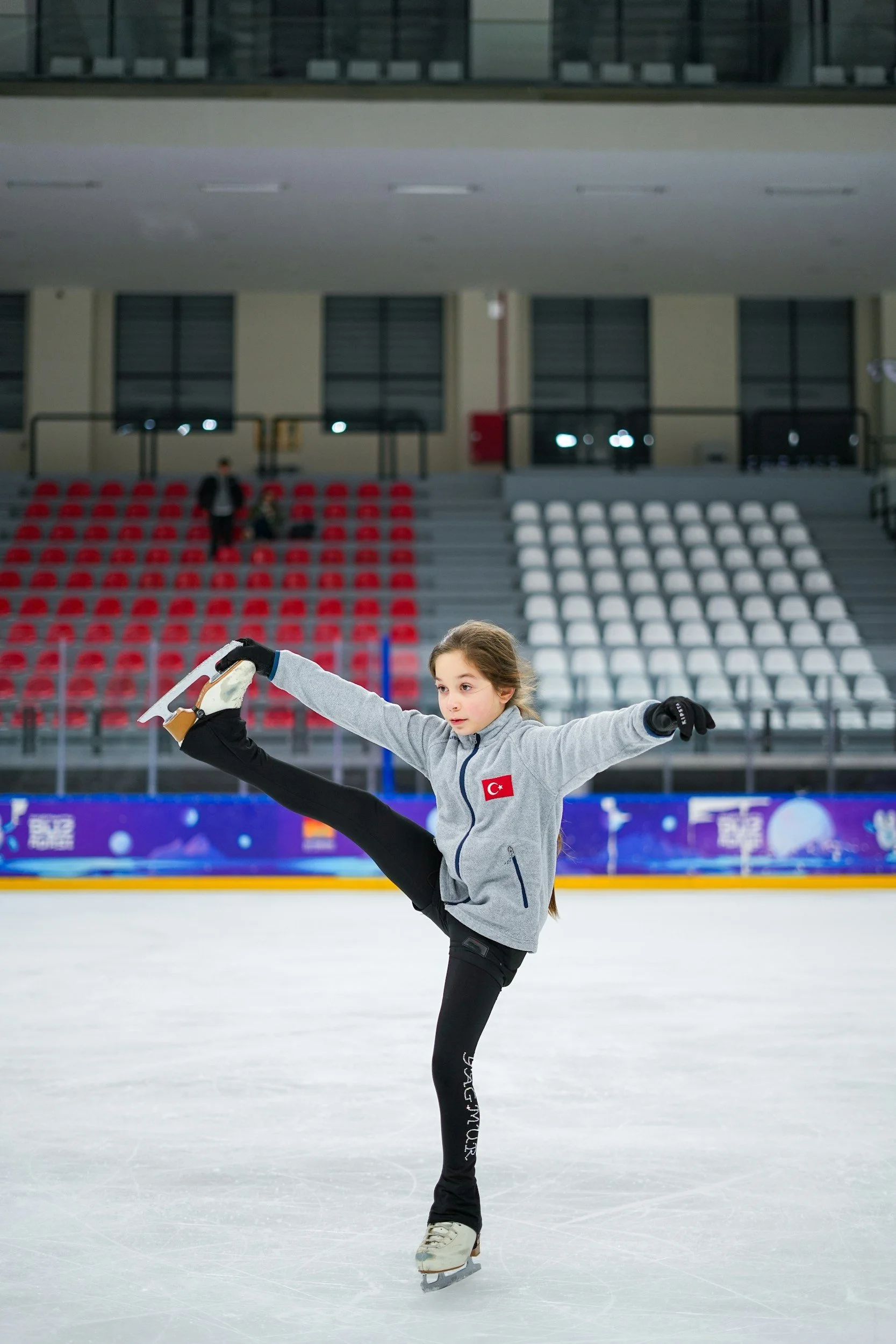 Young girl figure skating on an indoor ice rink, wearing a gray jacket with a Turkish flag patch and black pants, performing a move with one leg extended.