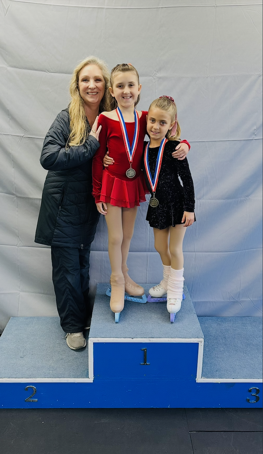 Two young female figure skaters with medals, standing on a winners' podium, alongside an adult woman, all smiling at the camera.