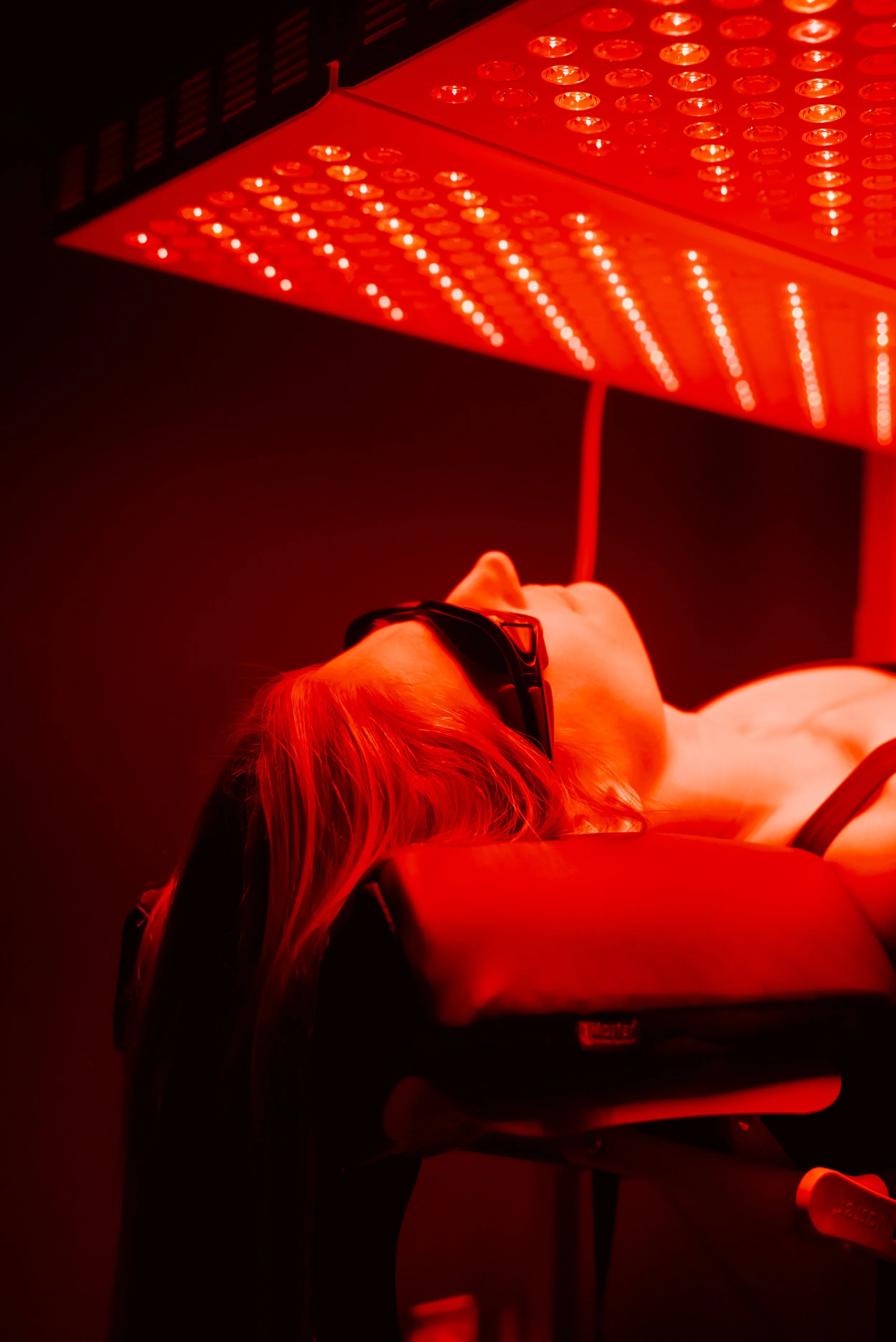 A woman in workout clothes stands next to a red-orange LED light panel, stretching her arms above her head in a fitness or gym setting.