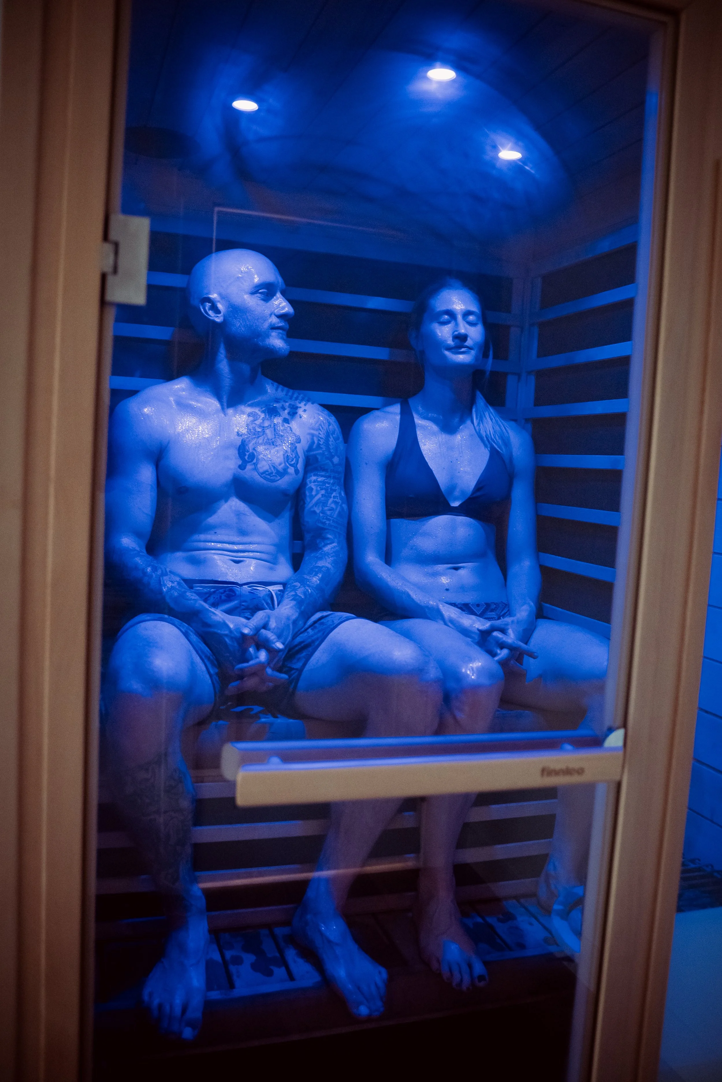 Interior of a wooden sauna with two benches, a slatted ceiling, and two black speakers mounted on the ceiling.