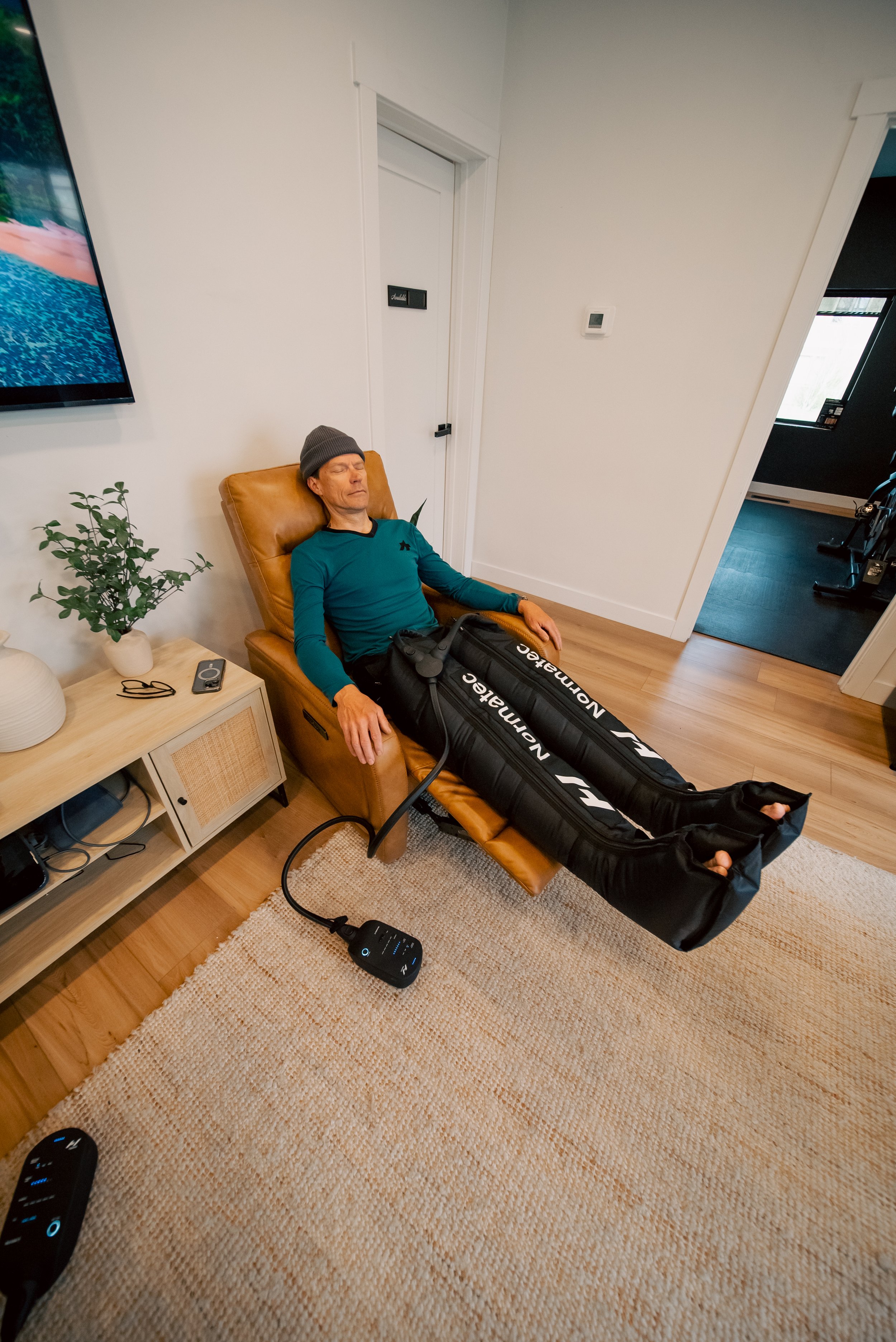 A person lying on a black leather sofa with a gray long sleeve shirt and gray pants. They are resting receiving Normatec compression therapy on both legs from feet to thighs.