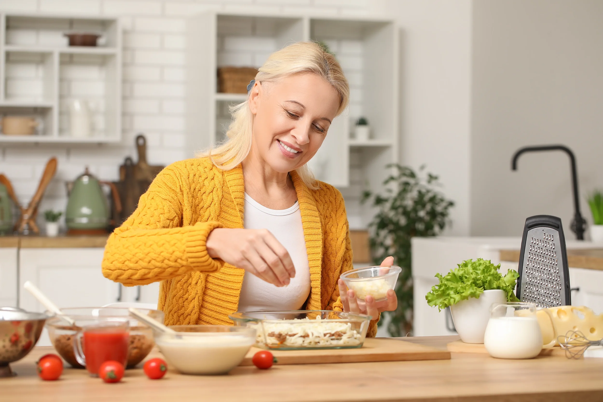 A woman preparing food in a kitchen with white cabinetry, wearing a yellow sweater and a white shirt, smiling while adding ingredients to a dish on a wooden counter.