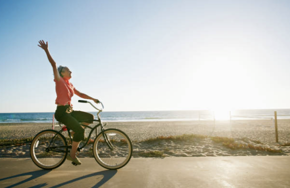 Woman riding a bicycle along a beach at sunset, raising her hand.
