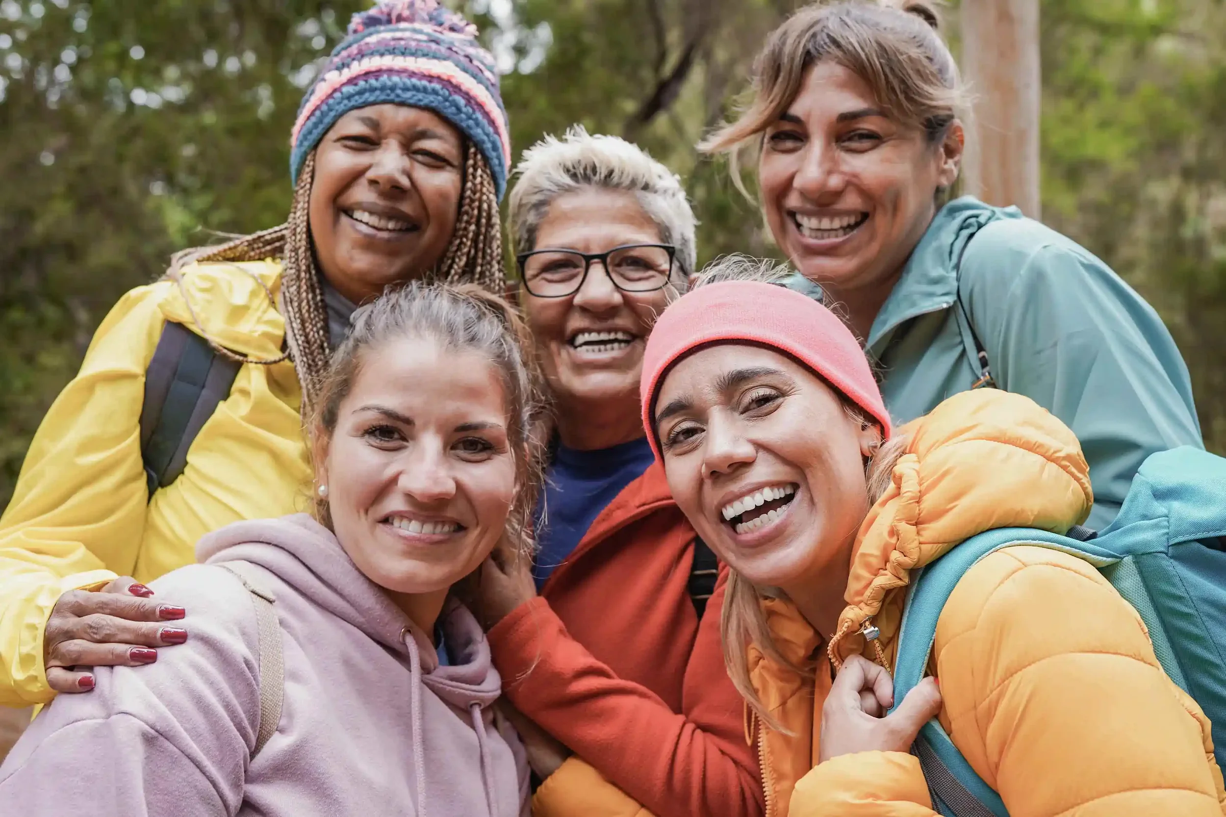 Group of six women smiling and posing outdoors in a forest, wearing colorful jackets and backpacks.